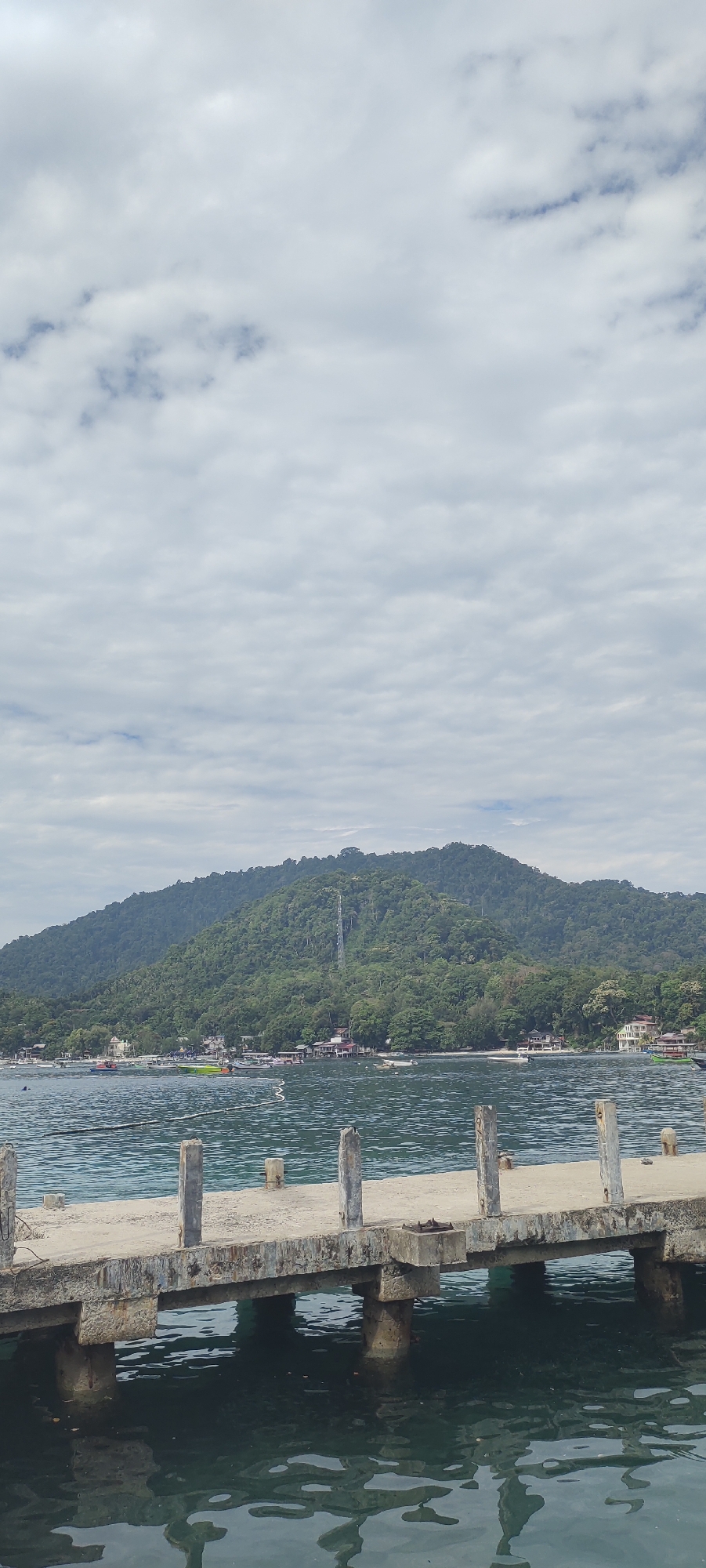 The image shows a view of the coast in Sabang, Indonesia, from a concrete pier extending into the water. The pier is weathered and shows signs of age, with rusty metal fixtures and concrete pillars supporting its structure. The water is calm and reflects the sky and the pier. In the background, a lush green, tree-covered mountain rises from the coastline, with a thin waterfall cascading down its side. Along the base of the mountain, a line of buildings, likely houses and shops, is visible, suggesting a settlement. Several small boats are anchored or moored in the bay in front of the settlement. The sky is overcast with scattered clouds, creating a diffused light over the scene. The overall impression is a tropical coastal landscape with signs of human habitation and natural beauty.