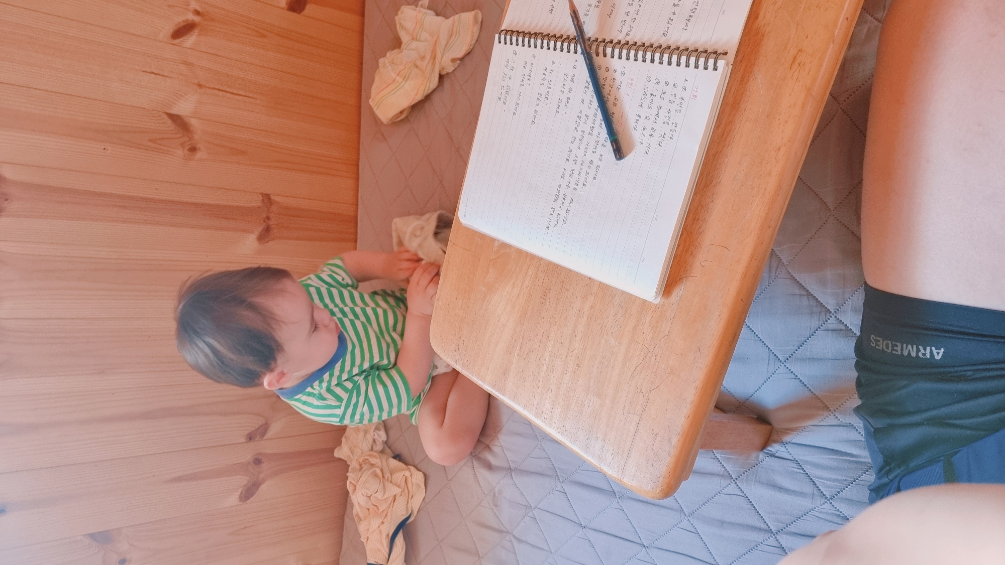 Here's a detailed description of the image:
**Setting:** The photo appears to be taken inside a small, possibly enclosed wooden structure with wooden walls. A blue-grey quilted mattress serves as the base.
**People:** A young child, presumably a baby or toddler, is sitting on the mattress. They're wearing a green and blue striped shirt. Partially visible is an adult person wearing dark colored sportswear.
**Objects:**
* A small, light wooden table is positioned in the scene.
* A spiral-bound notebook and a pencil lie on the table, filled with what appears to be handwritten text.
* A couple of folded cloths, likely cloths/wipes are near the child.
**Actions:** The child seems to be playing with the cloths. The presence of the notebook and pencil suggests someone has been studying or writing.
**Interaction:** The child is occupied with cloths, and it is possible that the adult person is studying Korean.
**Contextual Note:** OCR output mentions geographical place "Dunnae-myeon" and various phrases. It's highly possible that the notebook contains Korean language learning notes.
**Overall Impression:** The scene depicts a casual, intimate moment, possibly someone multitasking study or work with childcare within a limited space.