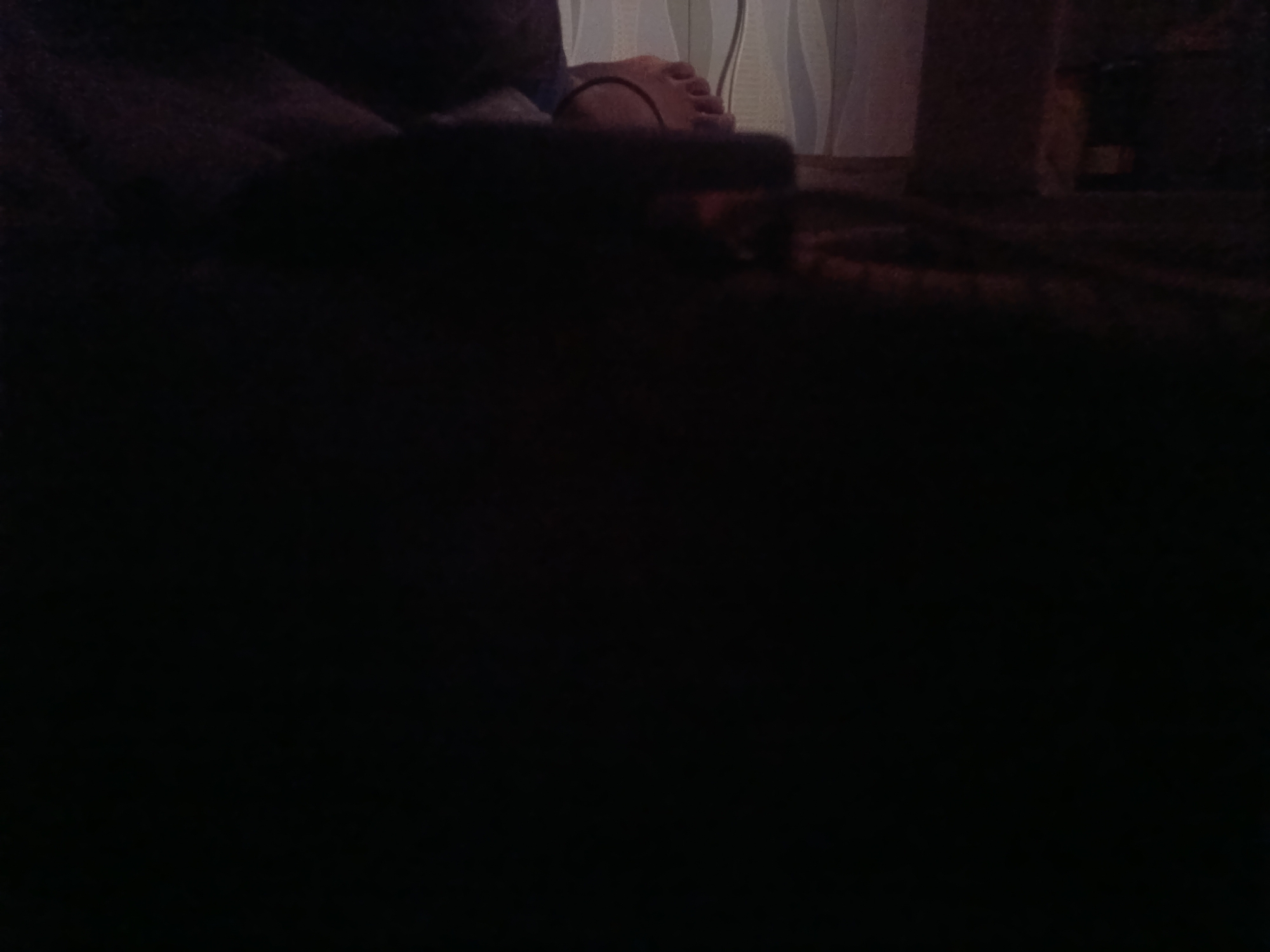 An overexposed, dark image shows a person's left foot with a thin black band around the ankle. The foot is resting on a dark surface. In the background, sheer curtains are visible, suggesting an indoor setting near a window. The overall lighting is very low, with significant noise and blown-out highlights.
