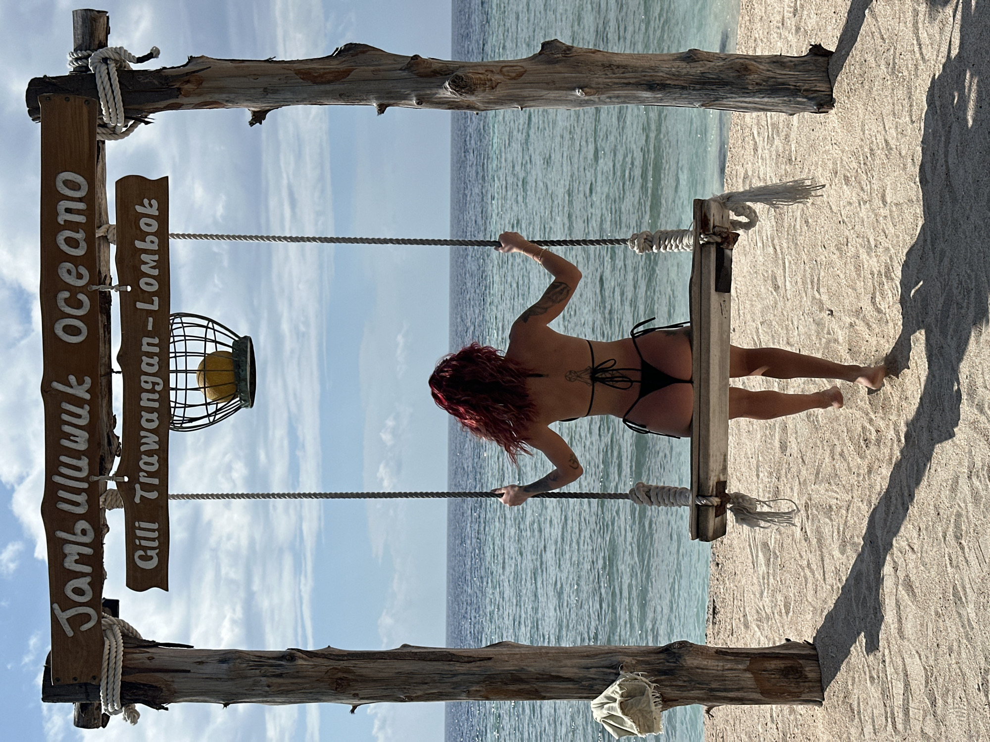 A woman in a black bikini stands on a swing set located on a beach in Gili Trawangan, Lombok, Indonesia. The swing set frame is made of weathered wood, with a sign reading "Jambuluwuk Oceano Gili Trawangan-Lombok" hanging on the left side. A decorative lamp is suspended from the frame. The woman has red hair and tattoos on her back and arms. The scene shows the beach, ocean, and a blue sky with some clouds in the background.