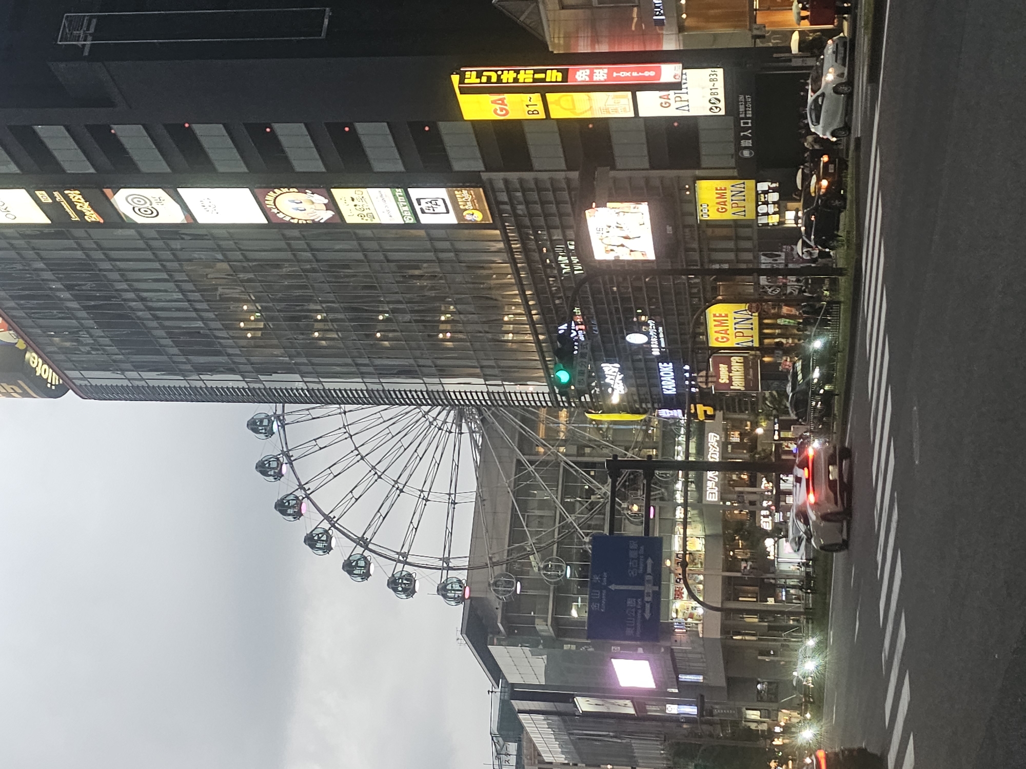 The image shows a street scene in Nagoya, Japan, at dusk. A large Ferris wheel is visible in the background, as well as a tall building with many illuminated signs. Cars are driving on the street, and pedestrians are walking on the sidewalks. The overall atmosphere is urban and lively. Specifically, the image appears to have been taken near the Sakae district in Nagoya, known for its entertainment and shopping venues, including a Ferris wheel.