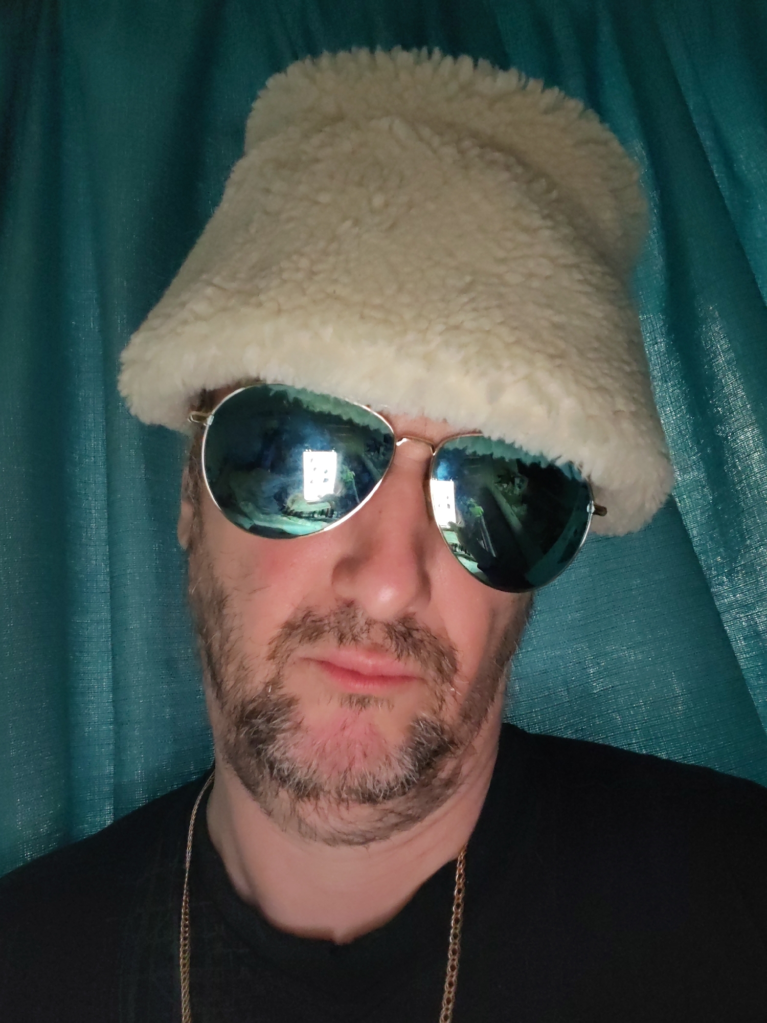 A man wearing a white, fuzzy hat and aviator sunglasses poses for a close-up shot. He has a beard and mustache, and a gold chain is visible around his neck. He is wearing a black t-shirt. The background is a teal-colored, textured material. The reflection in his sunglasses shows what appears to be an interior space with lighting fixtures. The photograph does not provide sufficient information to confirm the location in the city of Chaikovsky, Russia, or any activities or interactions.