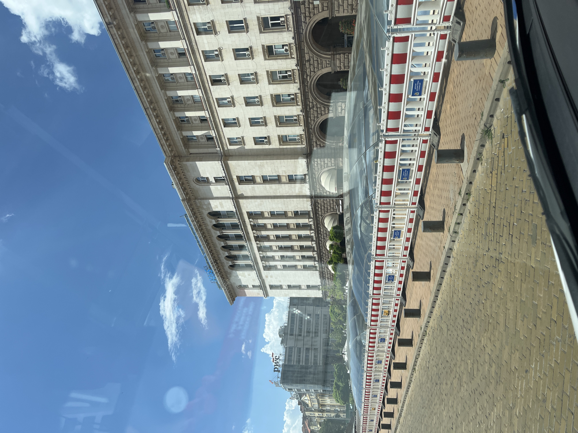 The image captures a street scene, likely in a city, viewed from a vehicle. The perspective is rotated, making the ground appear to run vertically along the right side of the frame. Dominating the left side of the image is the facade of a large, ornate building with multiple stories and many windows. The building appears to be constructed of light-colored stone or concrete, with decorative molding and arched details.
In front of the building, a section of what looks like a construction barrier or scaffolding is visible. This barrier consists of white metal frames with alternating red and white striped panels. It is covered by a transparent, possibly glass, enclosure, creating a reflective surface.
Further into the scene, another building is visible, this one appearing more modern and constructed of grey concrete. A logo featuring the letters "PWC" is clearly visible on this building. The sky above is a bright blue, dotted with fluffy white clouds, suggesting it is daytime with fair weather.
The scene suggests an urban environment where construction or renovation is taking place. The presence of the PWC logo might hint at a commercial or business district. The overall impression is one of a bustling, possibly sunny day in a city.