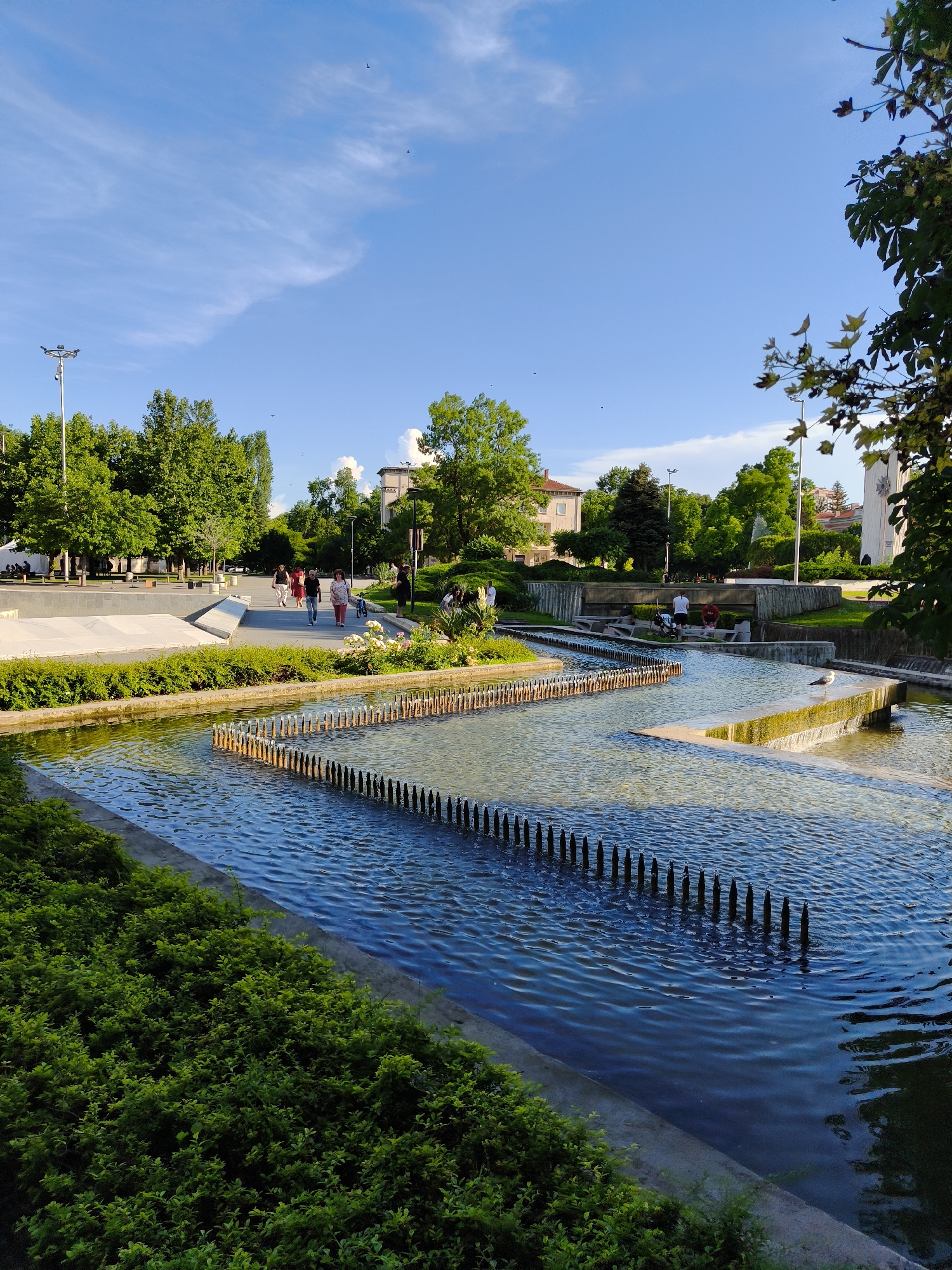 The image depicts a view of the central fountain complex in the city of Pleven, Bulgaria, during daylight. The foreground features a body of water with a tiered, zigzagging design, lined with numerous dark, upright posts. Lush green bushes border the water on the left side. In the midground, a paved walkway is visible, where several people are walking. Some are dressed in casual attire, with one person walking a small dog. Behind the walkway, there are more tiered water features and concrete structures, with a fountain spraying water in the background. The background is filled with mature green trees and buildings, including a light-colored structure with a tower on the right. The sky is blue with scattered white clouds and some wispy cirrus clouds, and a few birds are visible in the sky. The overall scene suggests a public park or city square during a pleasant day.