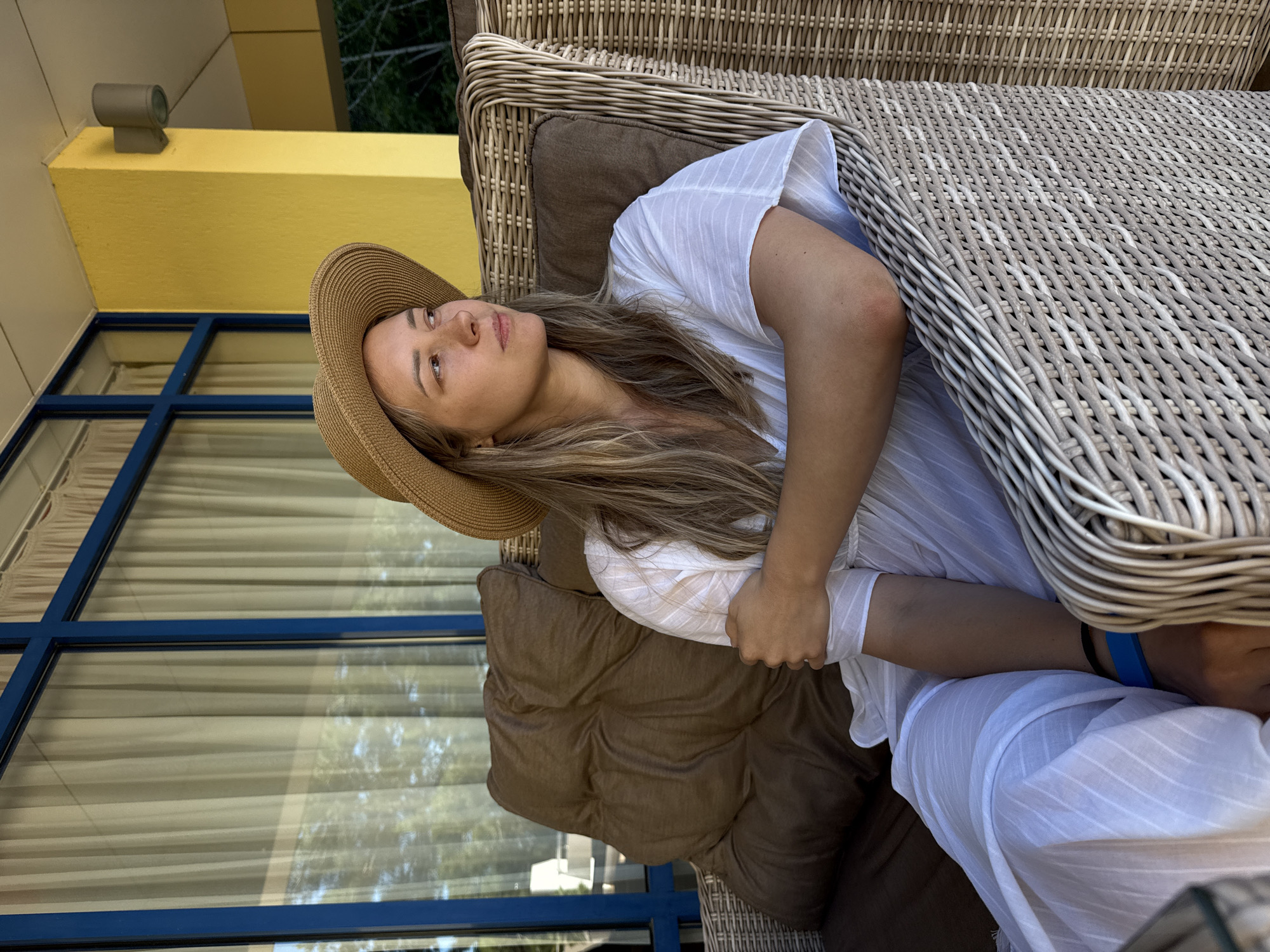 A woman wearing a straw hat and a white dress is seated on a wicker chair, looking to her right. She has long, blonde hair and her arms are crossed. She is on a covered patio, with large windows and sheer curtains in the background. A bright yellow pillar is visible to her right. The location is identified as Nebug, Russia.