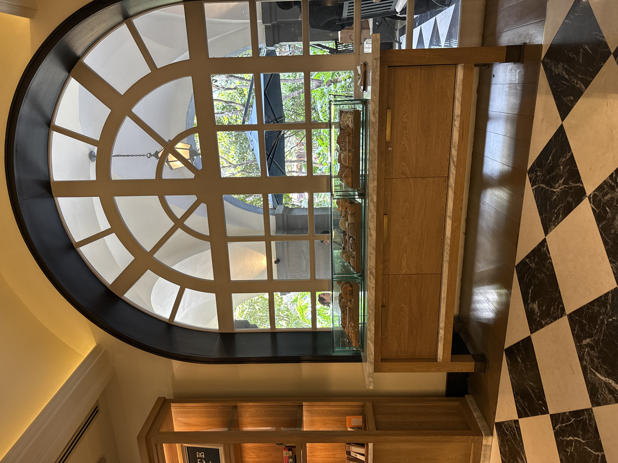 The image depicts an interior scene with a large, arched window divided into multiple panes. The window provides a view to the outside, showing trees and a building exterior. To the right of the window is a tall wooden cabinet with a glass display case on top, likely showcasing pastries. Below the window, a wooden bookshelf is visible, containing books and other items. The floor is covered in a black and white checkered pattern. The location is Mexico City, Mexico.