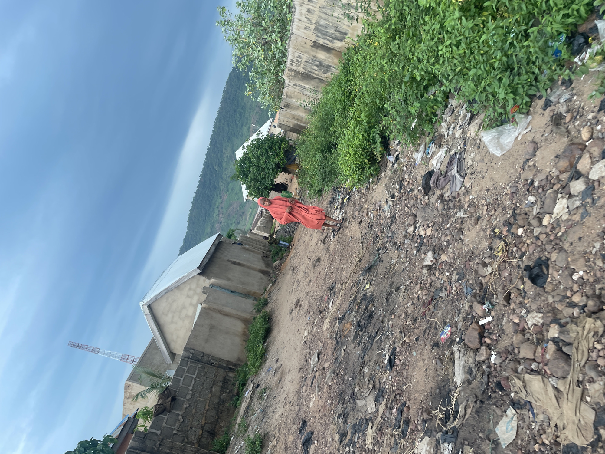 In Lokoja, Nigeria, a person wearing a bright orange religious garment walks on a dirt path. The path is littered with various debris, including rocks, plastic bags, and other refuse. To the left of the path are low-lying buildings constructed from concrete blocks, some with corrugated metal roofs. A tall telecommunications tower is visible in the distance above the buildings. To the right of the path, a concrete wall runs along the property line, overgrown with lush green vegetation. Beyond the wall, more buildings are visible, with a green hillside forming the backdrop under a cloudy sky.
