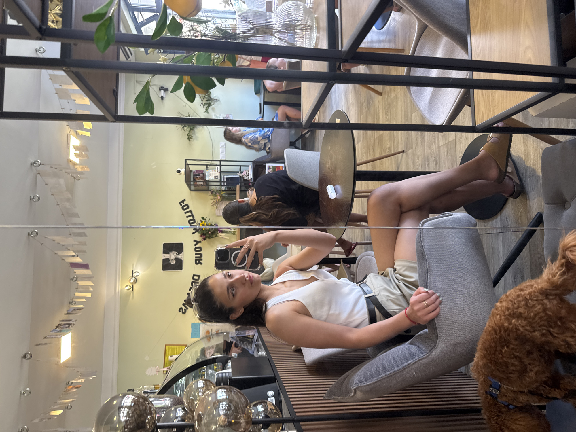 This detailed image captures an indoor scene within a cafe located in Budva, Montenegro, primarily through a mirror reflection. The central subject is a young adult female, seated in a grey upholstered armchair, taking a selfie with a black smartphone held in her right hand. Her dark hair is pulled back into a high ponytail, and she is wearing a white ribbed v-neck sleeveless top, khaki shorts with a black belt, a gold ring on her left ring finger, a red string bracelet on her left wrist, and light blue nail polish. Her feet are clad in tan open-toe slides.
The mirror reflects the cafe's interior, featuring light pastel green walls and a white ceiling adorned with a garland of small, rectangular paper decorations strung across the upper perimeter. Text on the back wall reads "FOLLOW YOUR DREAMS" in reverse, positioned below a black-and-white framed image depicting a person holding a martini glass. Lighting fixtures are visible on the ceiling and walls.
Various tables and chairs furnish the space, including round wooden tables with black bases and other grey upholstered chairs similar to the subject's. A dark, slatted wooden counter or wall section is visible behind the main subject's reflection, featuring a glass display case and several large, reflective silver spherical decorations. Towards the right, a dark metal shelving unit with wooden shelves holds books, small plants, and a white security camera.
Other patrons are visible in the reflection: a woman with dark hair seen from behind at a table to the left, another woman in a blue and white patterned dress further back, and a child's head peeking over a table to the far right. Green foliage and plants are interspersed throughout the cafe, including some with yellow, lemon-like fruit. A clear, ribbed glass vase sits on a table, and a small white cylindrical object is also present on another table.
In the foreground, outside the mirror's reflection, the lower half of the main subject's legs and feet, including her sandals, are visible. Adjacent to her chair on the floor, a small, curly-haired brown dog, possibly a poodle, wearing a blue collar, faces away from the camera. The flooring appears to be light-colored wood or laminate.