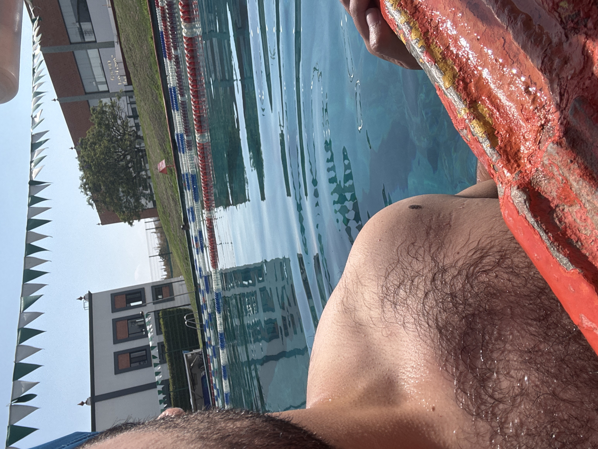 A man is in a swimming pool, holding onto a red and yellow life preserver. The pool has lane markers with red, white, and blue floats. Beyond the pool are buildings, a tree, green space, and green and white pennant flags. The location is San Andrés Cholula, Mexico.
