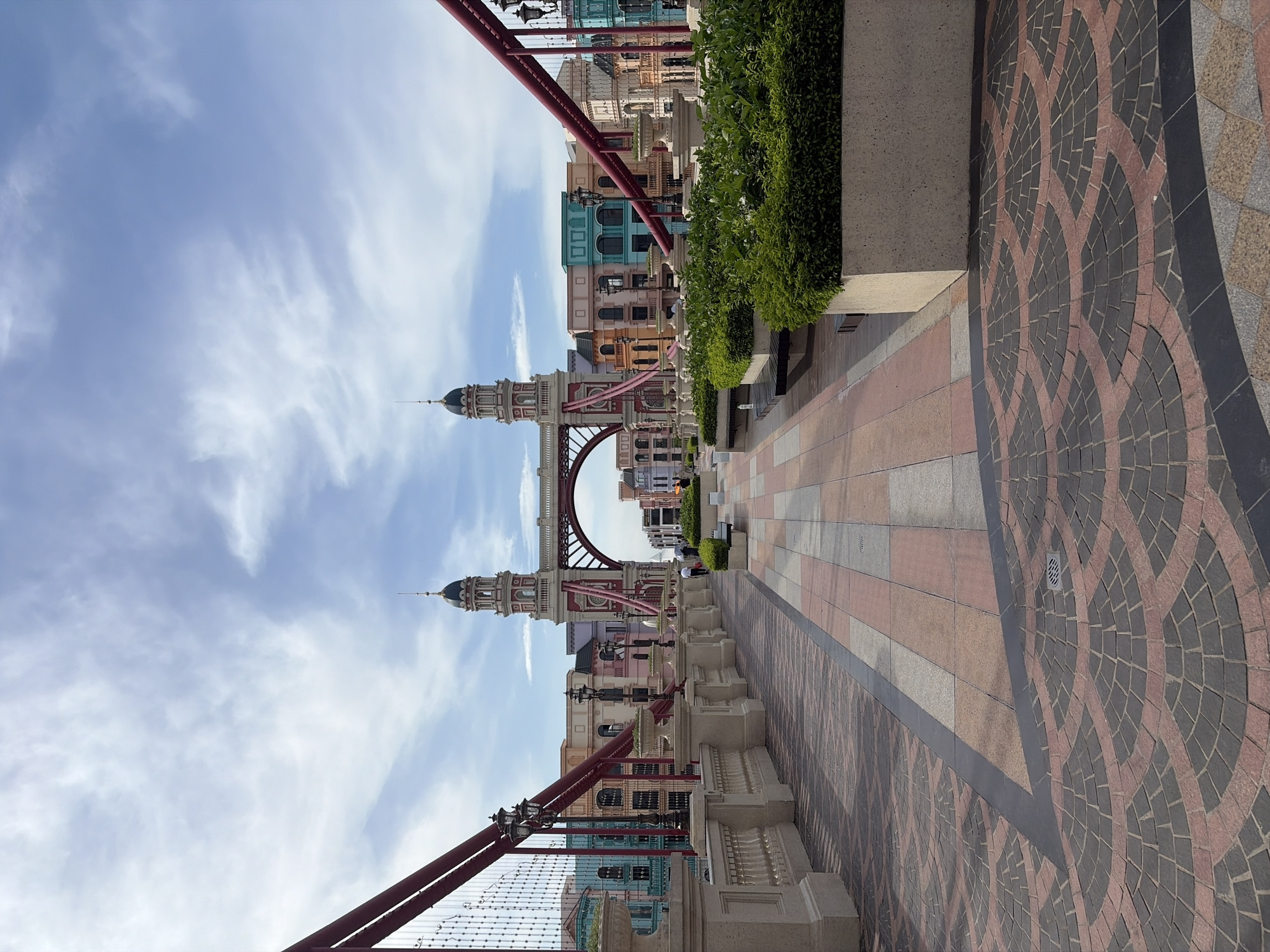 The image shows a bridge with a decorative railing and archway, leading to a row of colorful buildings. The bridge is paved with patterned tiles, and there are planters with green bushes along the sides. Two lampposts are visible on the bridge railing. In the background, the buildings have distinct architectural features, including arched windows and colorful facades in shades of blue, pink, orange, and cream. The sky is partly cloudy. There are two individuals walking on the bridge in the distance. The location is the city of Nghĩa Trụ Commune, Vietnam.