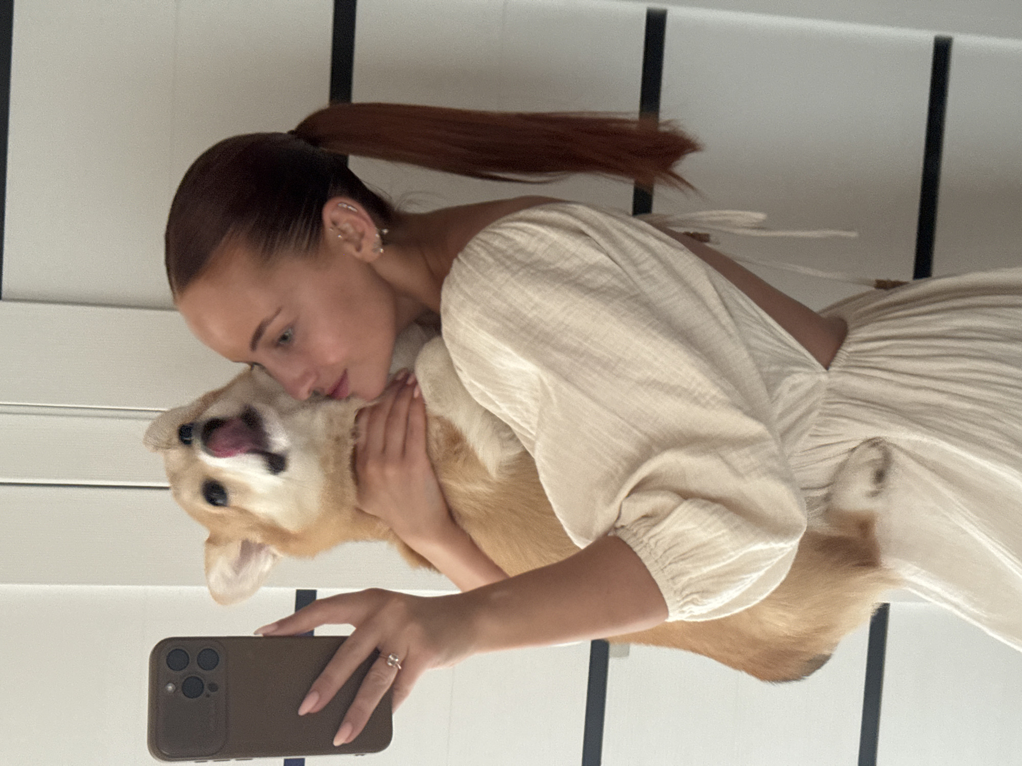 A woman with red hair pulled back in a high ponytail is embracing a corgi dog. She is holding a brown cell phone and appears to be taking a photo. The woman is wearing a light-colored dress with ruffled sleeves. The dog has its mouth open and tongue visible. The background features white walls with black vertical stripes. The specific location in the city of Chaikovsky, Russia is not discernible from the image.