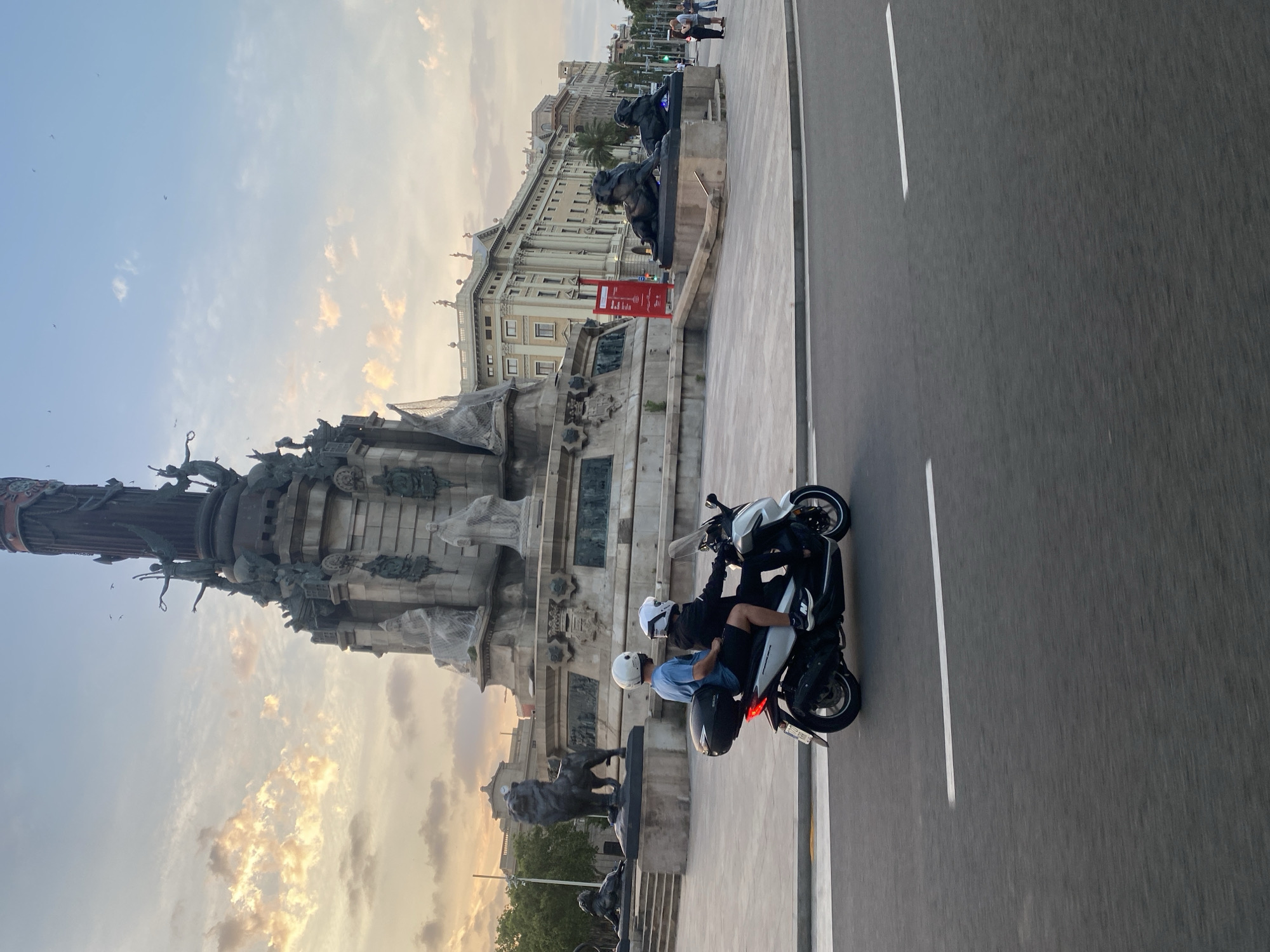 In Barcelona, Spain, two individuals wearing white helmets ride a white scooter on a paved road. The scooter has a black storage box attached to the rear. The passenger, wearing a blue t-shirt and black shorts, sits behind the driver, who is wearing a black hoodie and shorts. They are positioned in the foreground, moving from left to right.
In the background, a large monument with statues and intricate carvings dominates the scene. The monument is situated in a wide plaza with stone paving. Several lion statues are visible around the base of the monument, and a grand building with classical architecture stands behind it. The sky is partly cloudy with hints of sunset colors, and birds are scattered across the sky. To the right of the monument, a red sign is mounted on a pole. Further in the background, several people are walking in small groups on a sidewalk. Trees and lampposts line the street. The overall scene suggests an urban environment during late afternoon or early evening.