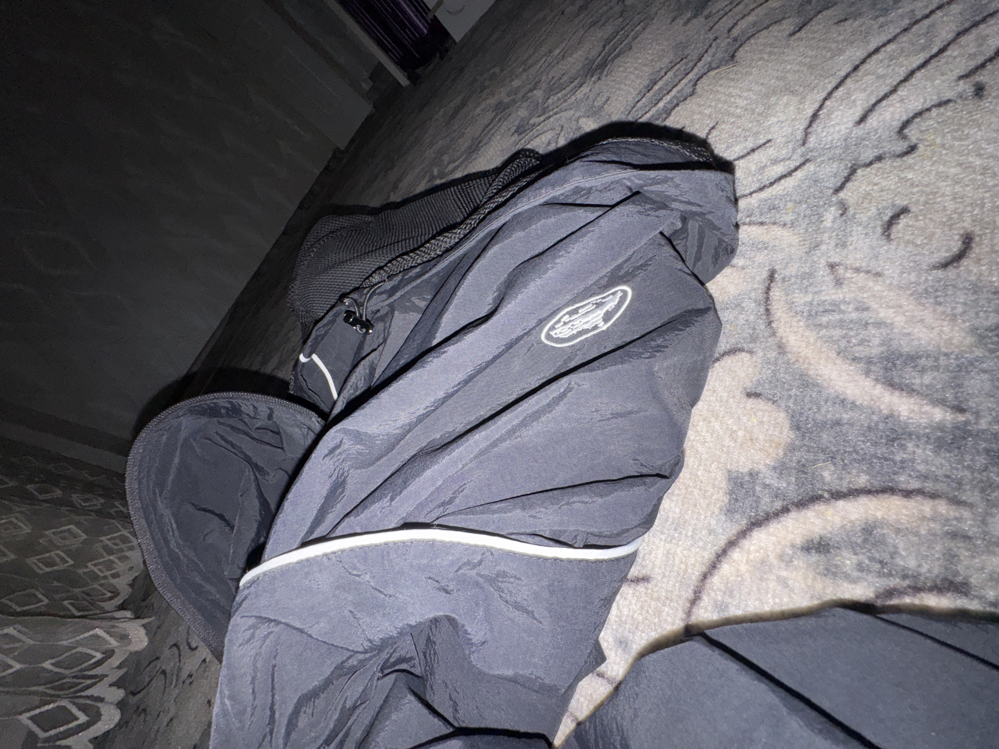 A black jacket with white trim and a circular logo is resting on a patterned blanket. A black backpack strap is visible. The background is dark and appears to be an interior space with a wall and what might be curtains or blinds. The scene is illuminated by artificial light, creating shadows. The location is identified as Kyzyl-Kyya, Kyrgyzstan.