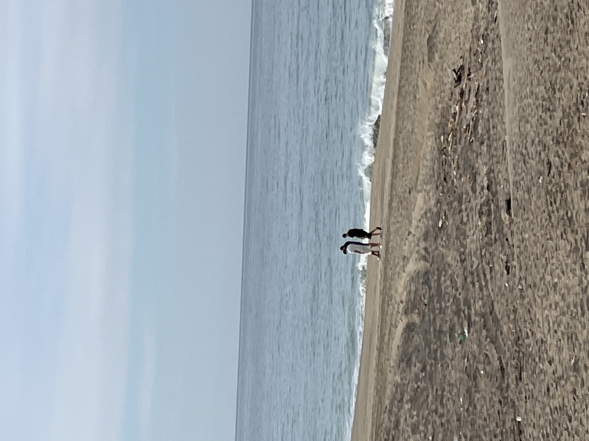 The image shows a beach in Alanya, Turkey. Two people are walking along the shoreline where the ocean meets the sand. The sky is overcast, and the water is calm. The sand appears to be dark. There is trash visible on the sand.