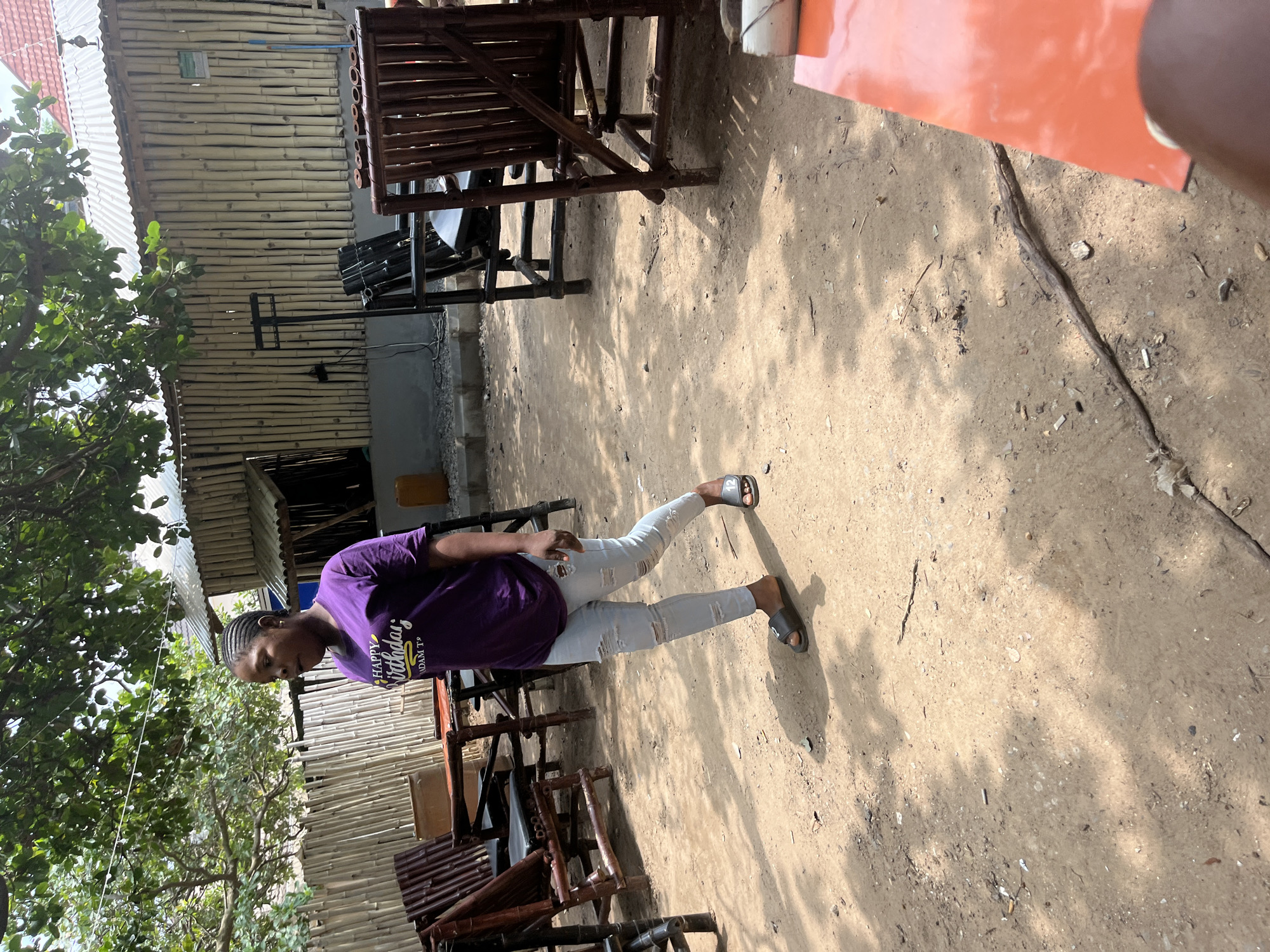 A person stands in a courtyard with a dirt ground. The person is wearing a purple t-shirt and light-colored ripped jeans. They are also wearing black sandals. The t-shirt has text that says "Happy Birthday". In the background, there are structures made of bamboo and corrugated metal roofing. There are also some wooden chairs and tables. Sunlight is casting shadows on the ground, suggesting it is daytime. The overall setting appears to be outdoors, possibly a market or a casual outdoor seating area. The person is looking down and to their right, with a slight smile on their face. The image conveys a casual and relaxed atmosphere.