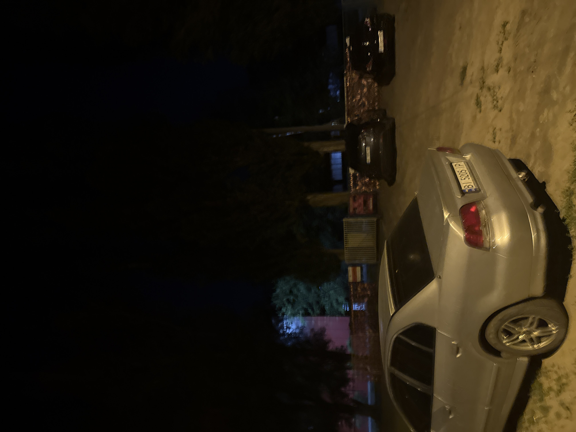 A silver sedan is parked on a dirt surface in front of a low brick building and trees at night. The car is angled towards the viewer's left, with its rear end closer to the camera. The license plate reads "BI 5205 IP". Two dark-colored cars are parked behind the silver sedan. In the background, a metal fence and more trees are visible. The scene appears to be in an outdoor parking area.