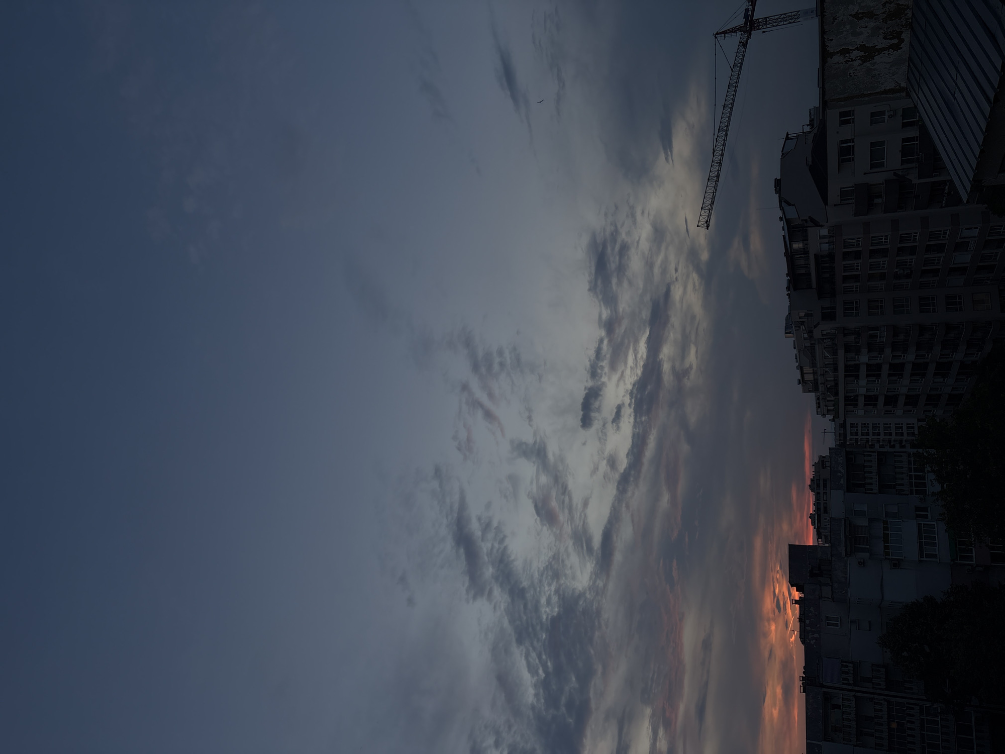 The image depicts a view of the city of Plovdiv, Bulgaria, at dusk. The sky is a deep blue transitioning to lighter shades near the horizon, with scattered clouds. A dramatic sunset is visible on the left side of the image, casting orange and pink hues across the sky and illuminating the edges of the clouds. Silhouetted against the sky are several apartment buildings, characteristic of post-socialist architecture, with balconies and numerous windows.
On the right side of the frame, a large construction crane is prominent, its latticed structure extending across the upper portion of the image. The crane suggests ongoing development or construction within the city. The lower part of the image is dominated by the dark outlines of rooftops and the upper floors of buildings. The overall atmosphere is one of twilight, with the city lights beginning to appear or still not yet fully visible. No people are discernible in the image. The scene captures a typical urban landscape at the end of the day, with the juxtaposition of residential structures and industrial construction equipment.