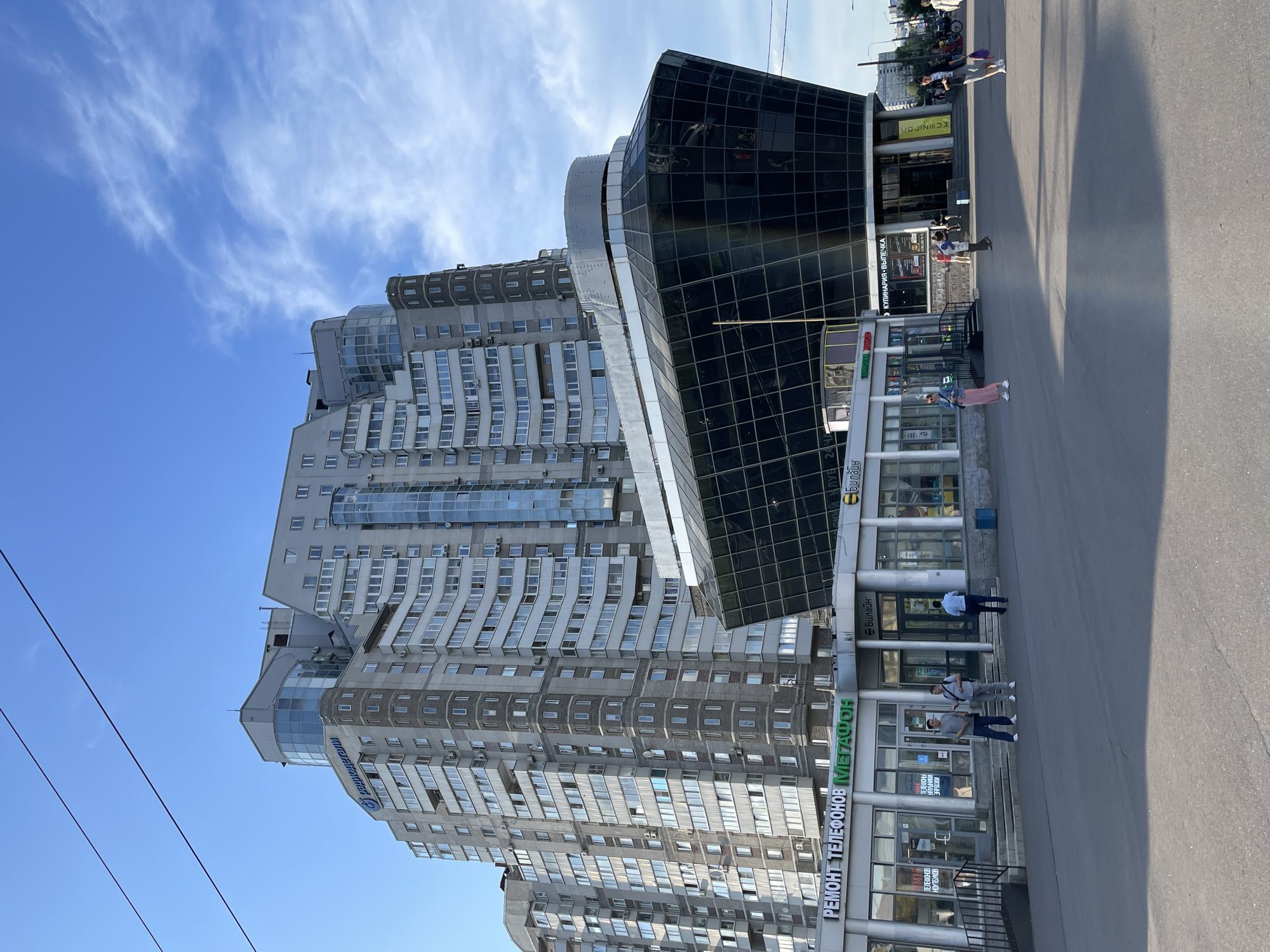 This is a full, eye-level, outdoor shot of a modern, multi-story building in Saint Petersburg, Russia, on a clear, sunny day with some wispy clouds. The building has a complex, angular design with a curved, dark-glass section at the lower front, resembling a ship's bow. Above this, the building is a lighter gray with many windows and balconies. A blue logo with Cyrillic text, reading "Радиодетали" (Radiodetal), is visible on the upper part of the building. At street level, there are several storefronts with signs, including "РЕМОНТ ТЕЛЕФОНОВ" (Phone Repair) and "Мегафон" (Megafon), and another with "КУЛИНАРИЯ" (Culinary) and "ВЫПЕЧКА" (Baking). Several people are walking on the pavement in front of the buildings. The scene is viewed from across a paved area, likely a sidewalk or plaza, with shadows cast by the buildings and overhead wires.