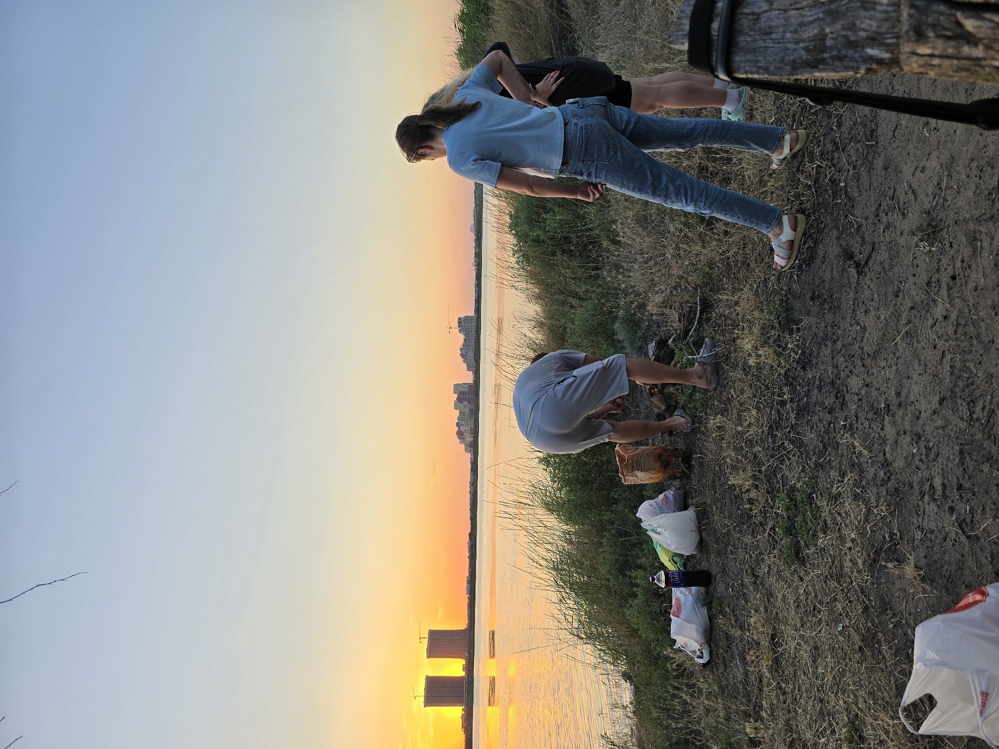 At dusk, a group of four individuals gathers near a body of water. A man in light gray shorts and a t-shirt bends over, seemingly tending to a fire pit, with a bag of charcoal and several plastic bags scattered on the ground beside him. To his right, two young women stand facing the water. One, wearing a blue t-shirt and jeans, looks out at the sunset, while the other, in a dark t-shirt and shorts, stands behind her. A fourth person, whose legs are visible, stands to the far left, also facing the water. The sun is setting over the water, casting a warm orange glow. In the distance, a city skyline with several tall buildings is visible. The scene is set on a grassy embankment, with dry grass and sparse vegetation covering the ground. The overall atmosphere suggests a casual outdoor gathering at sunset.