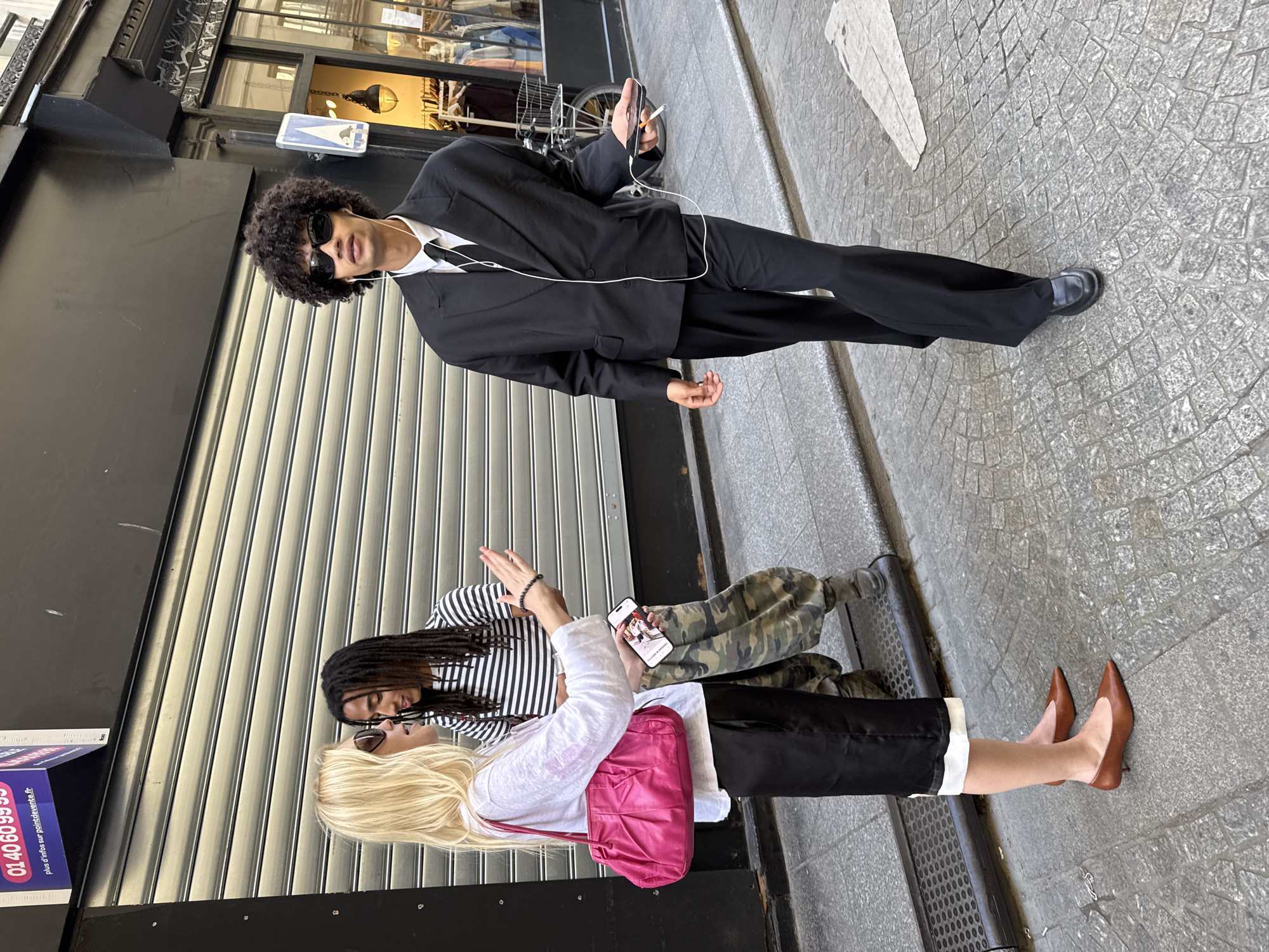 This street scene, set on a cobblestone sidewalk in Paris, France, captures three individuals engaged in distinct activities. In the foreground, a man with curly dark hair, wearing sunglasses, a black suit, white shirt, and black tie, stands confidently. He has white wired earbuds, holds a smartphone in his right hand, and a lit cigarette, with his left hand relaxed by his side.
Further down the street, two other people are engaged in conversation, leaning against or sitting near a closed shop shutter. One individual, with long dreadlocks, wears a black and white horizontal striped long-sleeved shirt and camouflage pants. They are facing the other person, appearing to listen. The second person has long blonde hair, wearing sunglasses on her head, a white long-sleeved top, black trousers with white cuffs, and brown pointed-toe high heels. She holds a smartphone in her left hand, which displays an image or video, and carries a vibrant pink shoulder bag.
The background features typical Parisian architecture, including shopfronts with closed grey metal roll-down shutters and ornate dark building facades with reflective windows. The scene is brightly lit by natural daylight, suggesting it is daytime. On a purple advertisement sign visible on the left, a phone number "01 40 60 99 95" is displayed along with French text "plus de mths sur demande ou renouvellement" (more months on demand or renewal). The overall mood is casual and observational, capturing everyday street life and contemporary fashion.