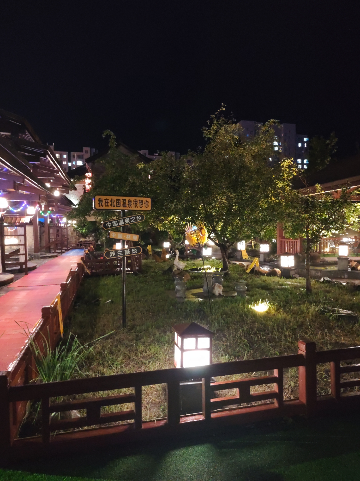 The image depicts an outdoor scene at night in Lin Dian Town, China. In the foreground, a red wooden fence runs horizontally across the frame, with a red pathway visible to the left. Beyond the fence and pathway, a grassy area is illuminated by several square, lantern-style lights. A signpost with multiple directional signs is situated in the grassy area. One sign reads "我在北国温泉很想你" (I miss you very much in the Northern Hot Springs). Another sign says "中国温泉之乡" (Hometown of Chinese Hot Springs). Other signs indicate "中国·林甸" (China·Lin Dian) and "养生基地" (Health Preservation Base).
In the middle ground, a tree with lush green leaves stands prominently, with more trees and foliage scattered throughout the area. A white goose figure is visible on the left side of the grassy area, near what appears to be a small pond. There are also some stone elements and small figurines present. In the background, modern apartment buildings with illuminated windows are visible against the dark night sky, indicating the proximity of a more urban setting to this recreational area. The overall ambiance suggests a tranquil, possibly tourist-oriented, location designed for relaxation and enjoyment of natural hot springs.