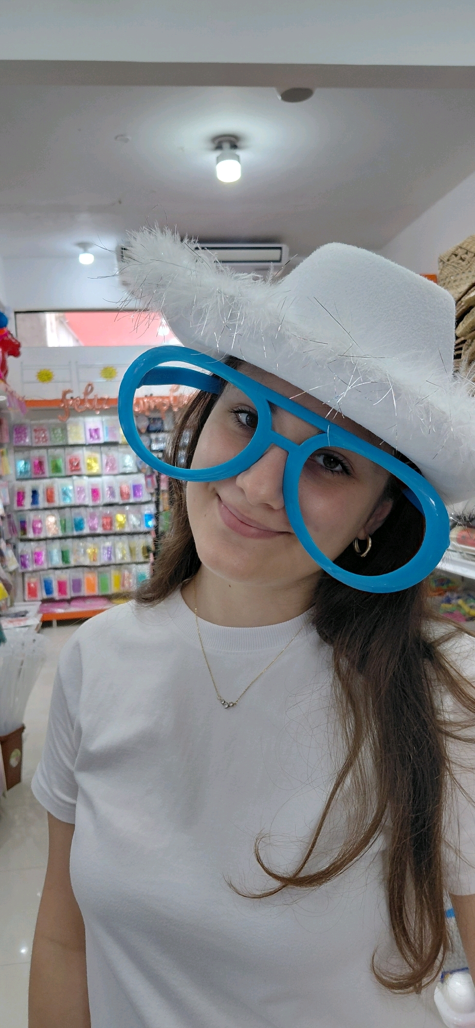 The image shows a young woman wearing a white feathered hat and oversized blue novelty glasses indoors. She is smiling and looking towards the camera. She is wearing a white t-shirt and a delicate gold necklace. The background appears to be a store with shelves filled with colorful items, possibly party supplies or accessories. The ceiling has light fixtures and an air conditioning unit. The scene is consistent with a retail environment in Encarnación, Paraguay, likely a party supply or costume shop given the props.