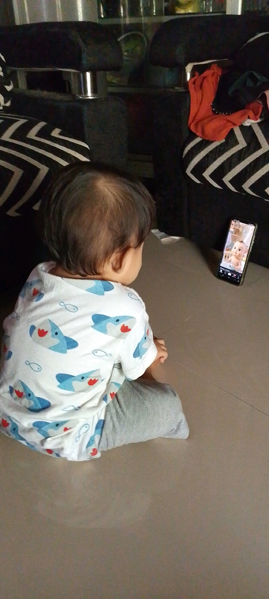 A rear view shows a young child sitting on a tiled floor, facing a smartphone. The child wears a white t-shirt with a pattern of blue sharks and grey pants. The smartphone, placed on the floor to the child's right, displays a video of a baby. In the background, a dark sofa with a black and white striped pattern is visible. The scene appears to be indoors, possibly a living room. The location is specified as Sindangrasa, Indonesia.