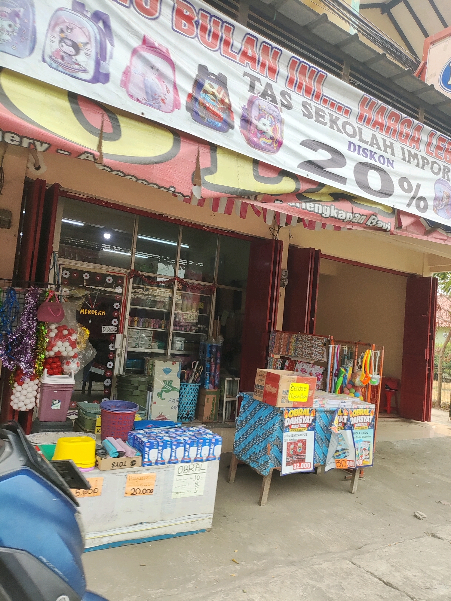 The image shows a street-level view of a shop in Wanajaya, Indonesia. A large banner across the top of the storefront displays a sale with text that includes "TAS SEKOLAH IMPOR" (Imported School Bags) and "DISKON 20%" (20% Discount). Below the banner, the shop's entrance is visible, with red folding doors. Inside, shelves are stocked with various items, and a display of merchandise is set up outside the shop. In the foreground, a blue motorcycle is parked, partially obscuring a white counter with items for sale, including boxes of what appear to be light bulbs. Signs on the counter indicate prices, such as "5.000", "20.000", and "32.000". The overall scene depicts a commercial area on a street.