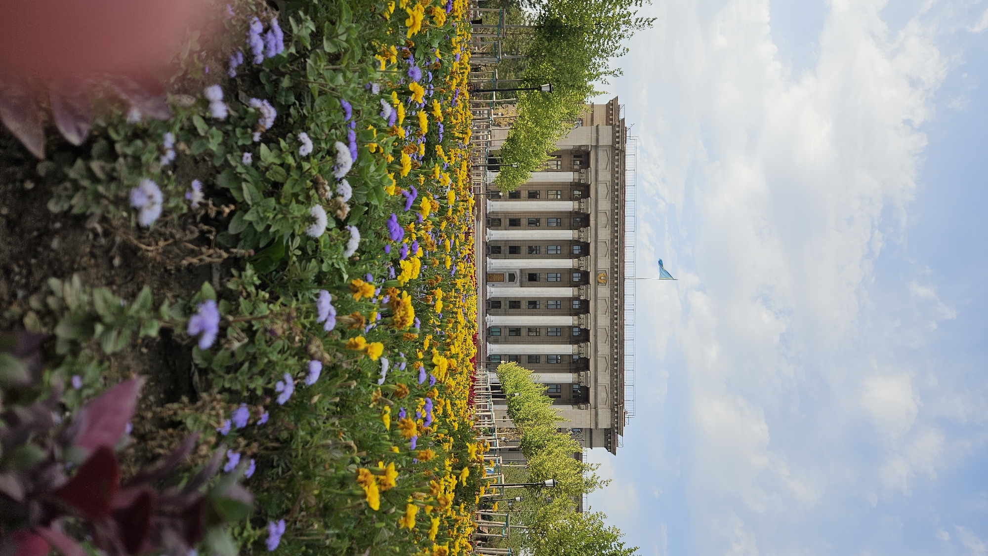 The image shows the facade of a large, neoclassical building with a row of tall columns. A light blue flag flies from the top of the building. In the foreground, a vibrant flower bed is filled with yellow and purple blossoms. The scene is set against a sky with scattered white clouds. This is the Republic Square in Almaty, Kazakhstan, featuring the former Presidential Palace, now housing the Kazakh National Pedagogical University.