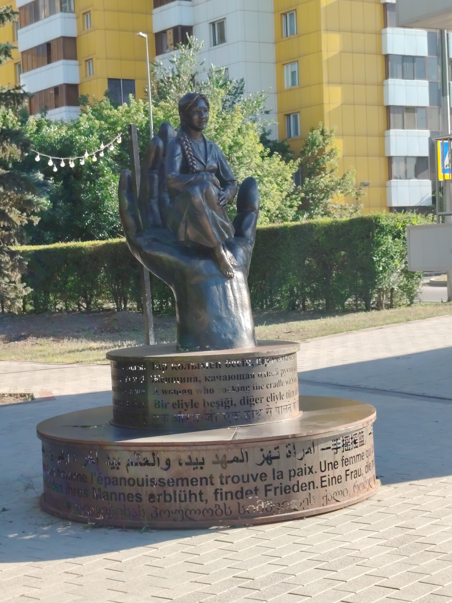 In the city of Astana, Kazakhstan, a bronze sculpture depicts a woman seated on a cupped hand, with her legs crossed. The sculpture is positioned outdoors, in front of a building with a yellow and white facade. The base of the sculpture features text inscribed in multiple languages, including Russian, English, Turkish, Hindi, Arabic, Chinese, French, German, and Georgian. The scene is illuminated by sunlight, casting shadows on the paved ground. Trees and hedges surround the sculpture, and a street light is visible behind it.