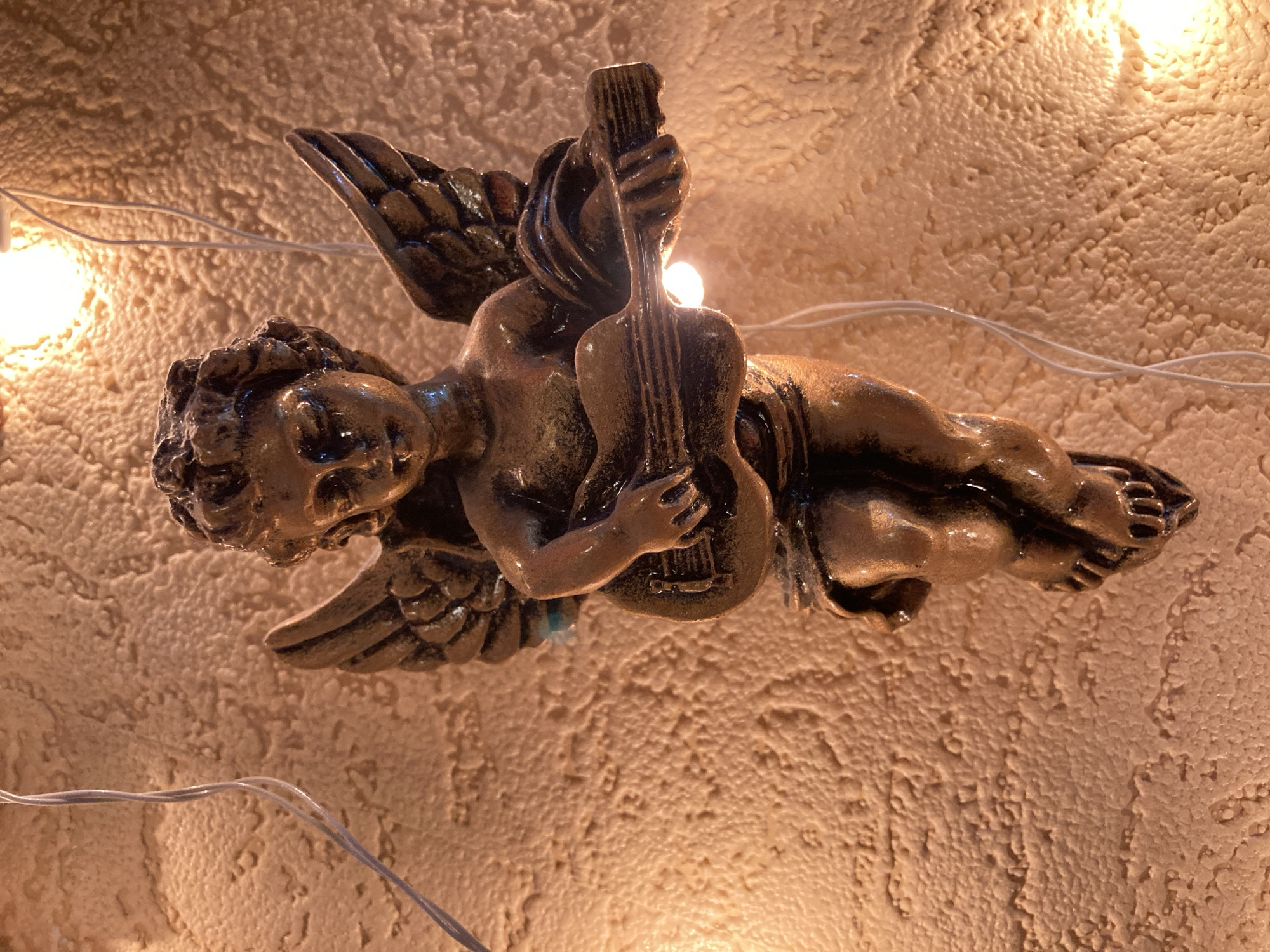 A bronze-colored statue of a cherub is affixed to a textured, light brown wall. The cherub is depicted in a reclining pose, holding a miniature guitar. Its wings are spread slightly behind it. Thin white wires are visible, attached to the wall and near the cherub, suggesting it might be part of a string of lights or decorations. The wall's surface has a rough, bumpy texture. Two bright lights are visible, one above and to the left of the cherub and another at the lower right of the image, casting a warm glow on the scene. The lighting creates strong highlights and shadows, emphasizing the contours of the statue and the wall.