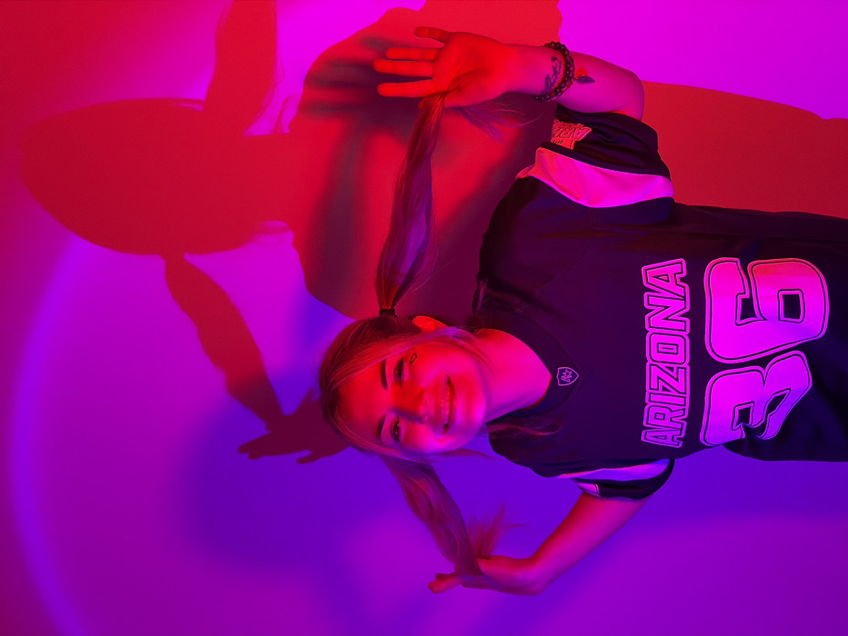 A female individual is depicted in an indoor setting within the city of Budva, Montenegro, illuminated by vibrant red and purple/magenta artificial light. The subject, a young woman with blonde hair styled into two pigtails, is smiling and looking directly at the viewer. She holds her left pigtail with her right hand and her right pigtail with her left hand. A small dark heart is visible on her right cheek. She is wearing a dark-colored sports jersey with "ARIZONA" and the number "36" printed in a bright pink/purple font on the front, along with a small shield-shaped emblem near the collar. A beaded bracelet is visible on her right wrist. Behind her, a prominent, elongated shadow of her figure, mirroring her pose, is cast in red light onto the wall, which transitions from purple/magenta on the left to red on the right. A distinct circular red light pattern is visible on the upper left portion of the wall.