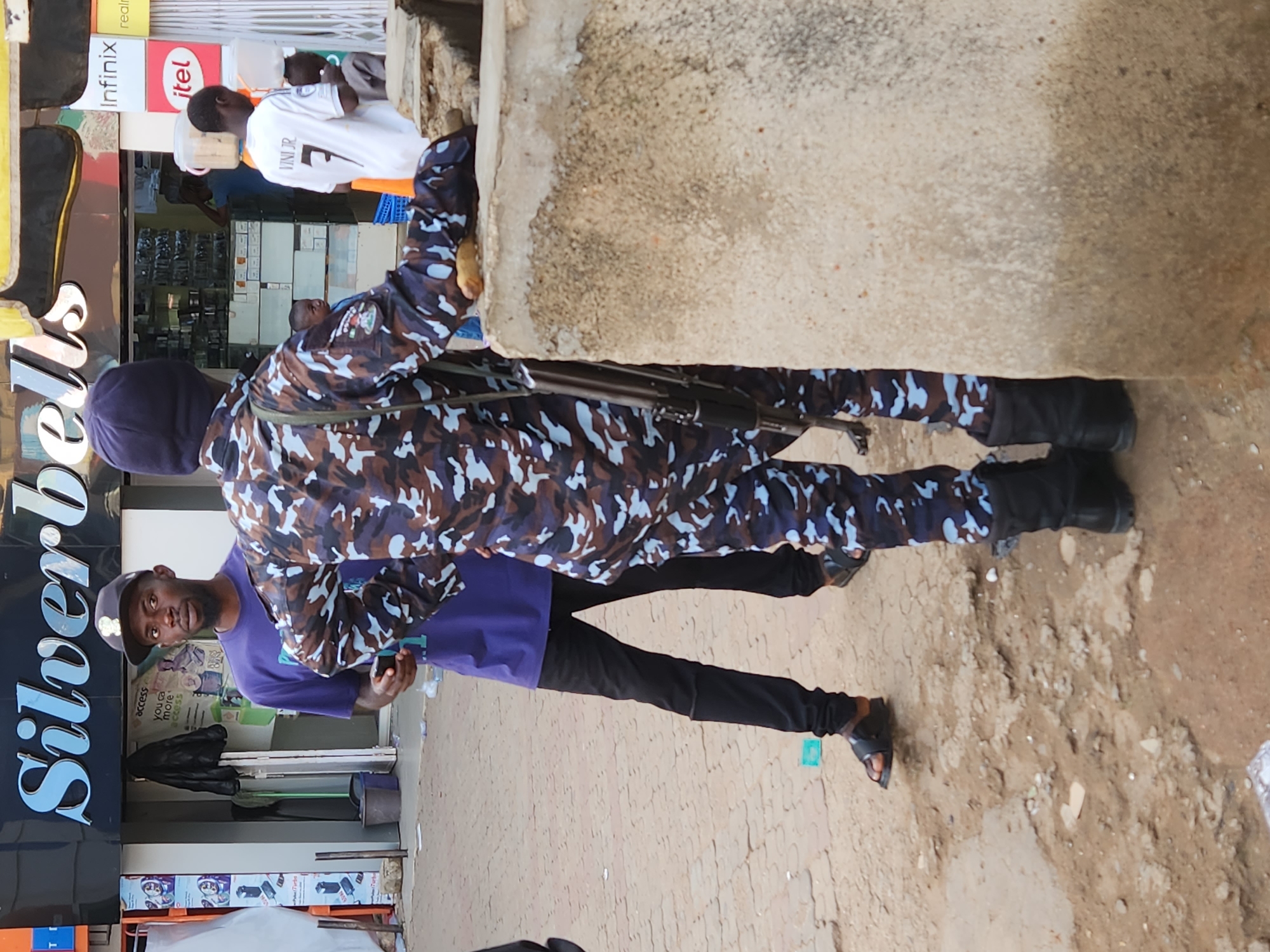 A medium-shot, rotated, outdoor image shows two men standing on a sidewalk in an urban area of Unknown, Unknown. The man in the foreground wears a dark cap, a purple shirt, and black pants and sandals. He is holding a mobile phone in his left hand and is looking directly at the camera. Behind him, a second man is dressed in a camouflage-patterned police uniform, complete with black boots and hat. He is carrying what appears to be a rifle. To the left, the signage for "Silverbells" is partially visible above what appears to be a storefront with glass displays of various items. To the right, a weathered concrete structure is situated, partially obscuring a man in a white jersey with the number "7" on it. Overhead, the signs for "Infinix" and "Itel" are seen. The background consists of a street view with other unidentified people.