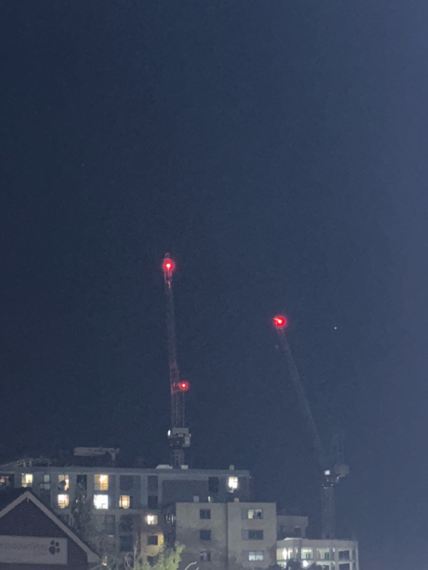 At night, two construction cranes are visible against a dark blue sky. The crane on the left has three red lights, two on its main structure and one at its apex. The crane on the right has one red light at its apex. Below the cranes, a multi-story building with lit windows is visible, indicating it is occupied. Part of a smaller building with a triangular roof is in the foreground on the left. The scene appears to be in Basildon, United Kingdom, given the context provided. There are no people or specific actions depicted.