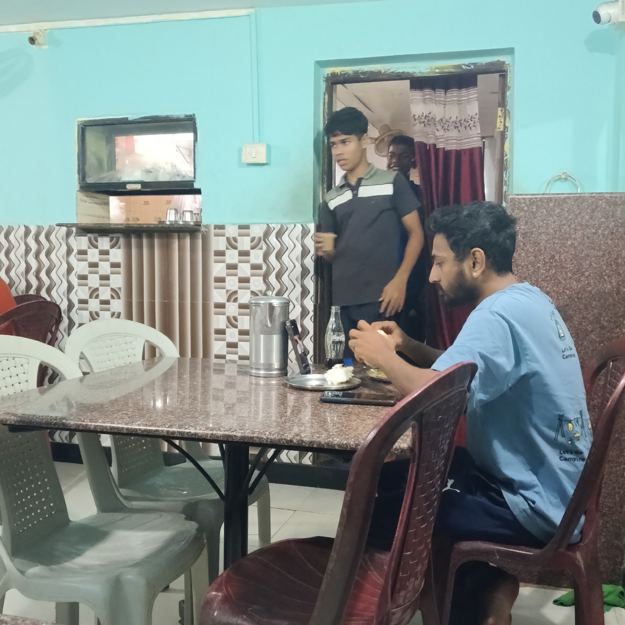 **Location:** Oxel, India
**Scene:** Interior of a modest dining area.
**People:**
* A man is seated at a table, eating. He has dark hair and a beard, and is wearing a light blue t-shirt with a camping-themed graphic on the back.
* Another younger man is standing near a doorway, holding a cup. He is wearing a dark gray shirt with a light gray stripe on the chest.
* A third man is partially visible through the doorway, behind the second man.
**Objects:**
* A table with a brown and gray patterned surface.
* Multiple white and burgundy plastic chairs are around the table.
* A stainless steel water pitcher and a bottle of Coca-Cola are on the table.
* A smartphone is lying on the table, near the seated man's plate.
* A metal plate with food is in front of the seated man.
* A security camera is mounted on the wall near a serving window, above decorative tiles and a shelf.
* A decorative curtain is visible through the doorway.
* A light switch is affixed to the wall.
* A towel ring is affixed to the wall.
**Actions:**
* The seated man is eating.
* The standing man is holding a cup.
**Environment:**
* Walls are painted a light blue color.
* The floor is tiled in a pattern of light and dark tiles.
* There is a serving window behind the table, with patterned tiles below.
* A doorway is visible in the background.