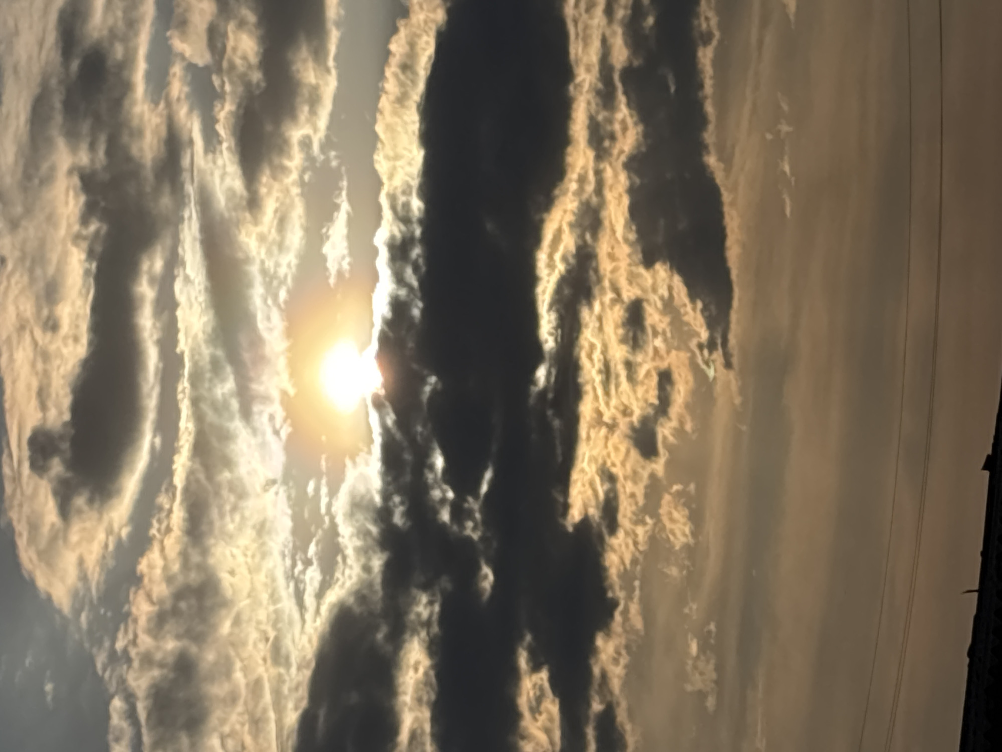 The image shows a sky filled with clouds, with the sun visible behind some of them. The clouds vary in color from light, almost white, to darker shades of gray. On the right side of the image, there are power lines and what appears to be a dark land area. The photo has a slightly warm tone due to the sunlight. The orientation of the image is rotated 90 degrees counter-clockwise.
The location and city are not discernible from the image.