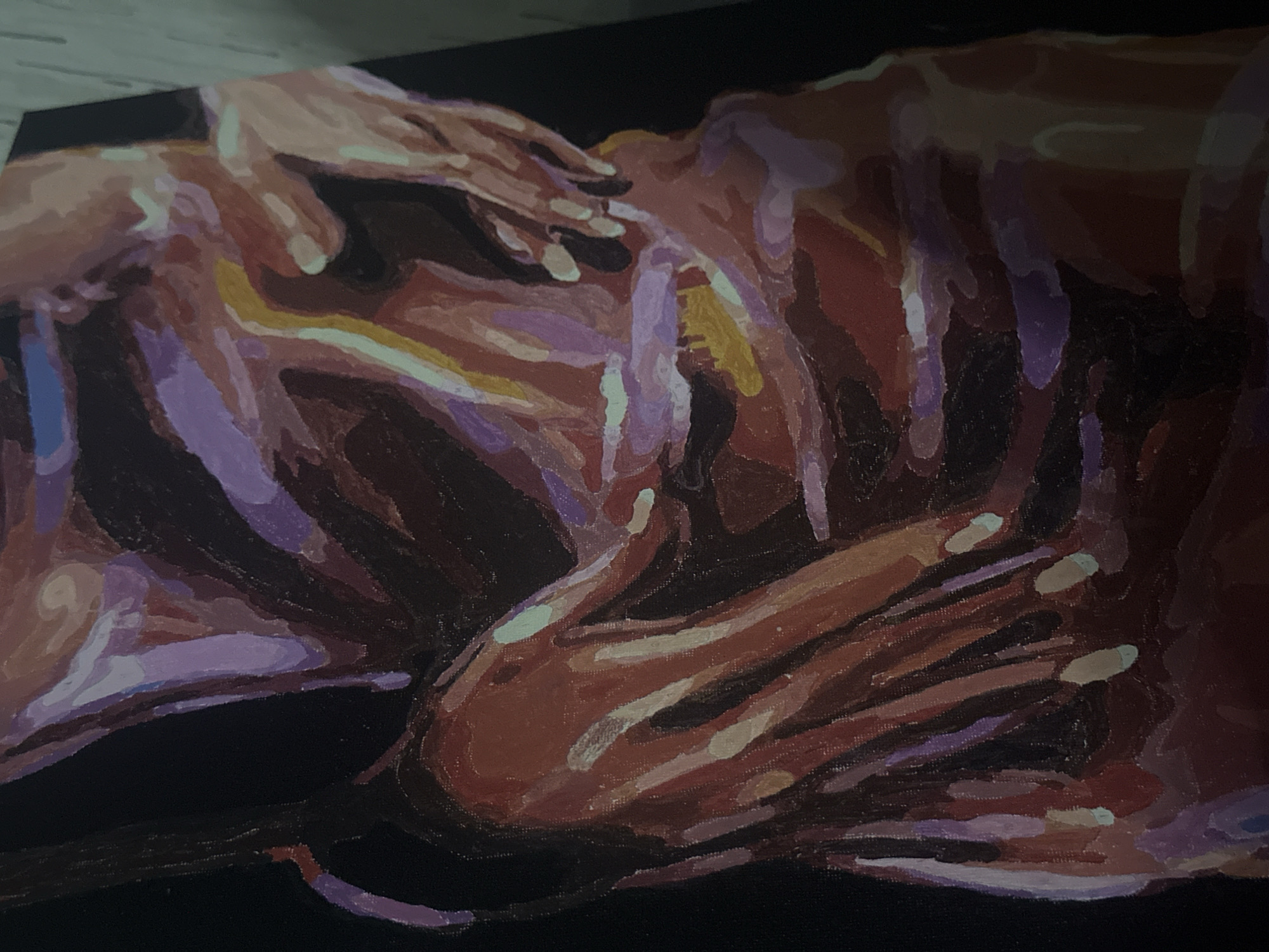 The image displays a close-up photograph of an expressive painting, likely created with oil or acrylic paints, depicting a human figure against a solid black background. The artwork focuses on the torso and limbs of a person in a reclined or supine position, rendered in a semi-abstract style. Distinct hands are visible; one hand rests on the upper back or shoulder area, while another hand is placed lower down, possibly on a leg or hip.
The painting employs a vibrant and contrasting color palette. Warm tones of red, orange, and brown are used for the body's primary forms, interspersed with prominent highlights and shadows rendered in cool shades of purple, lavender, light blue, and occasional touches of yellow and pale green. These bold colors and visible, confident brushstrokes create a textured and dynamic surface, emphasizing form and light in an impressionistic or expressionistic manner.
The figure's posture suggests a state of repose, rest, or quiet contemplation. No specific activity is taking place beyond this state of being. The setting within the painting is minimal, with the figure isolated against a dark void. The photograph of the painting shows it resting on a light-colored, possibly wooden, surface. There is no visible text in the image.