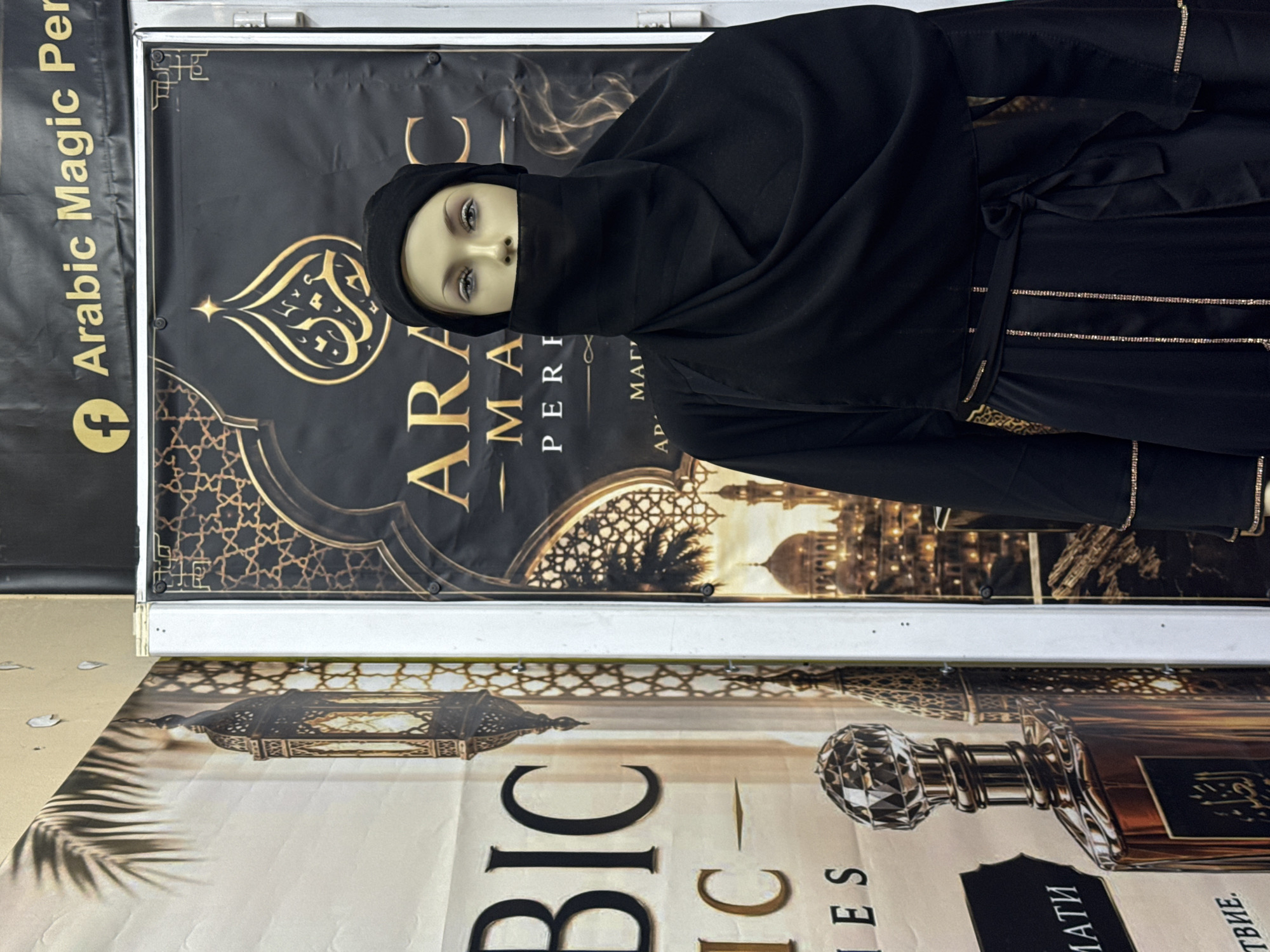 In Kiten, Bulgaria, a mannequin is dressed in traditional Islamic clothing, including a black hijab and niqab, standing in front of a display. The backdrop features a large banner with Arabic script and ornate patterns, along with images of lanterns and architectural elements characteristic of Islamic culture. To the left, another poster displays a perfume bottle and palm fronds. The overall scene suggests a retail environment showcasing cultural and cosmetic products.