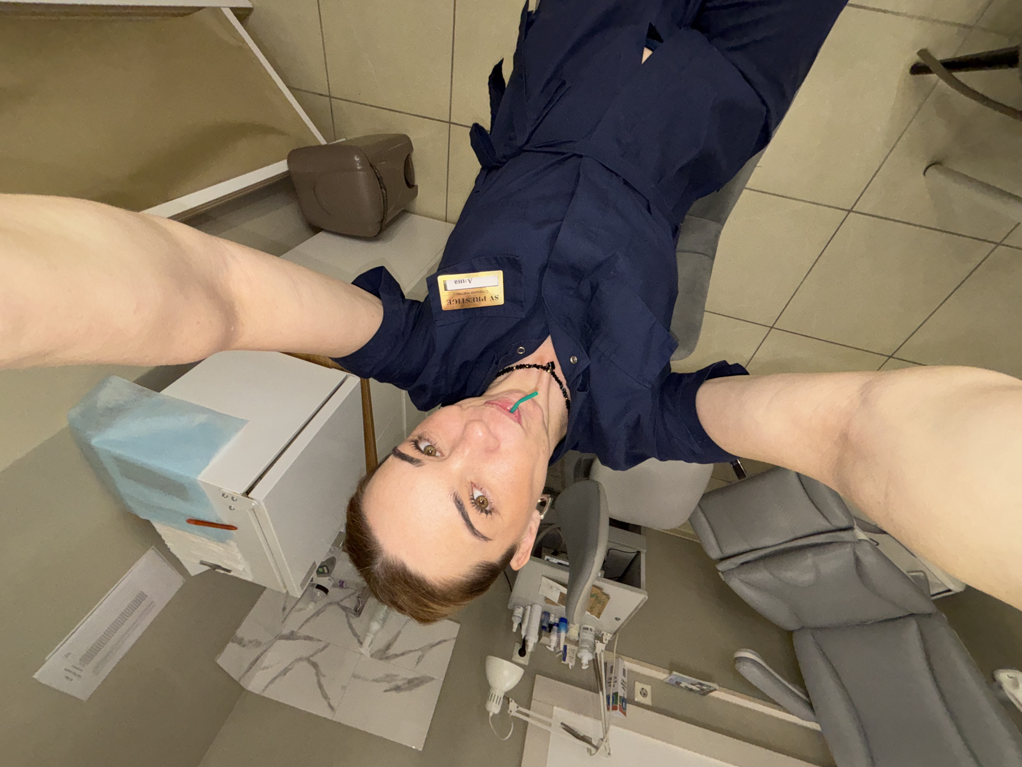 The image depicts a woman in a dark blue medical uniform in what appears to be a dental office. She is holding a green plastic item in her mouth and looking directly at the camera, which is angled from above. Her name tag reads "SV Prestige". The dental office contains equipment such as a dental chair, cabinets, a light, and tiled floors. The location is in Vsevolozhsk, Russia.