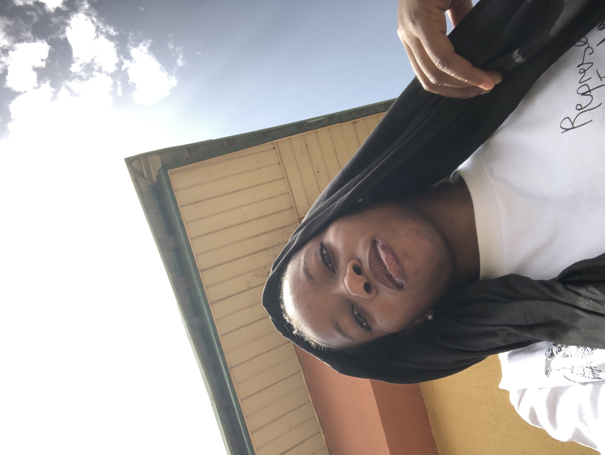 A low-angle, outdoor photograph shows a young, dark-skinned person with black fabric partially covering their head and a white t-shirt visible. The person is facing the camera and sticking out their tongue. The t-shirt has writing on it that is partially obscured. In the background, there is a building with light-colored horizontal siding and a dark green trim. Above the building, the sky is visible with some clouds. The photograph appears to be taken in a sunny environment. The location is Pindiga, Nigeria based on available data.