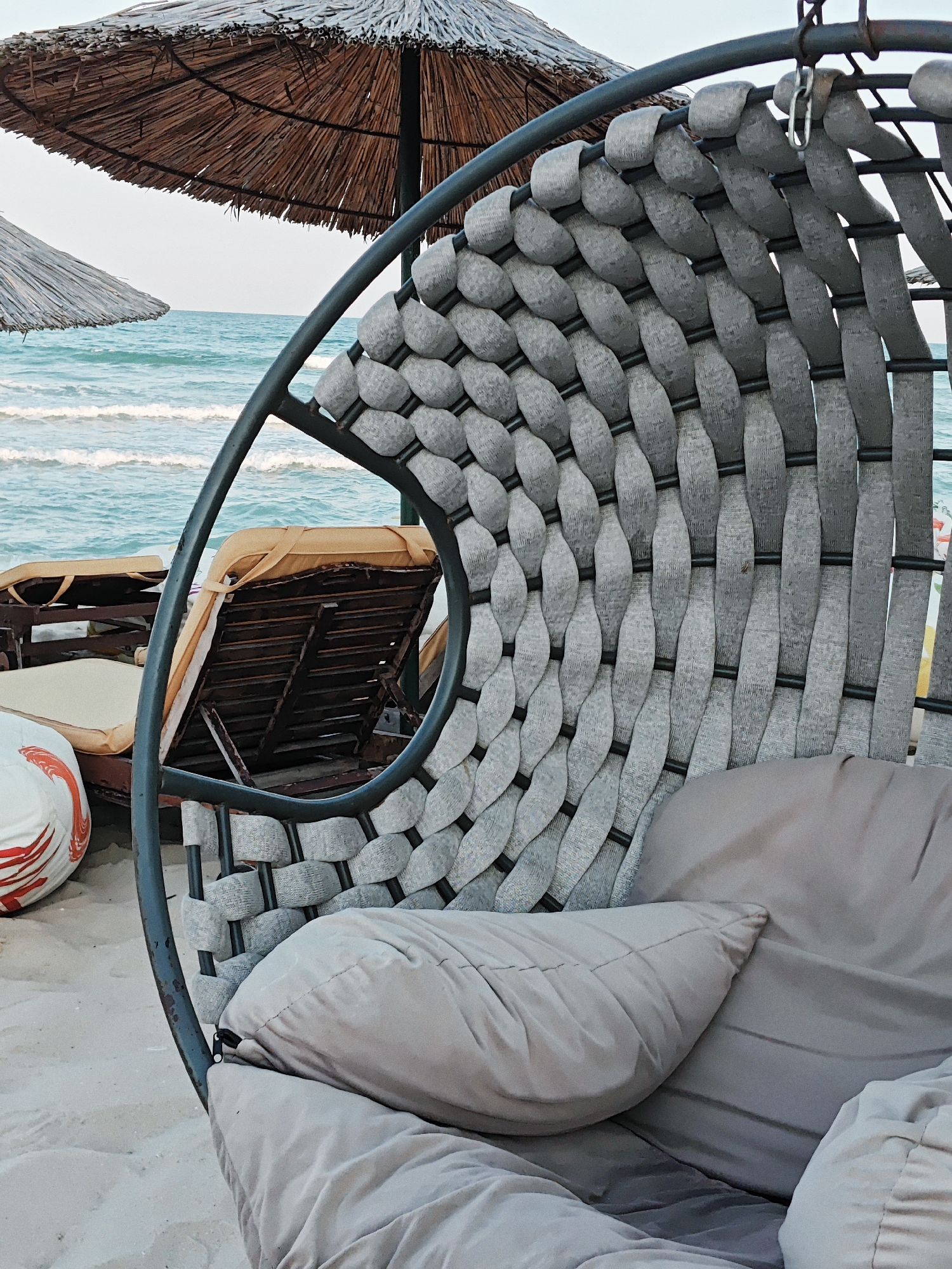 This is a medium shot, eye-level, outdoor photograph of a hanging swing chair on a sandy beach. The chair is made of grey woven fabric on a dark metal frame. It is cushioned with light grey pillows. To the left of the chair, out of focus, are two beach chairs with yellow cushions, and a large beach umbrella made of straw. Beyond the beach chairs and umbrella is the ocean with small waves and a clear sky. The overall scene depicts a relaxing beach setting. The location is Unknown, Unknown.