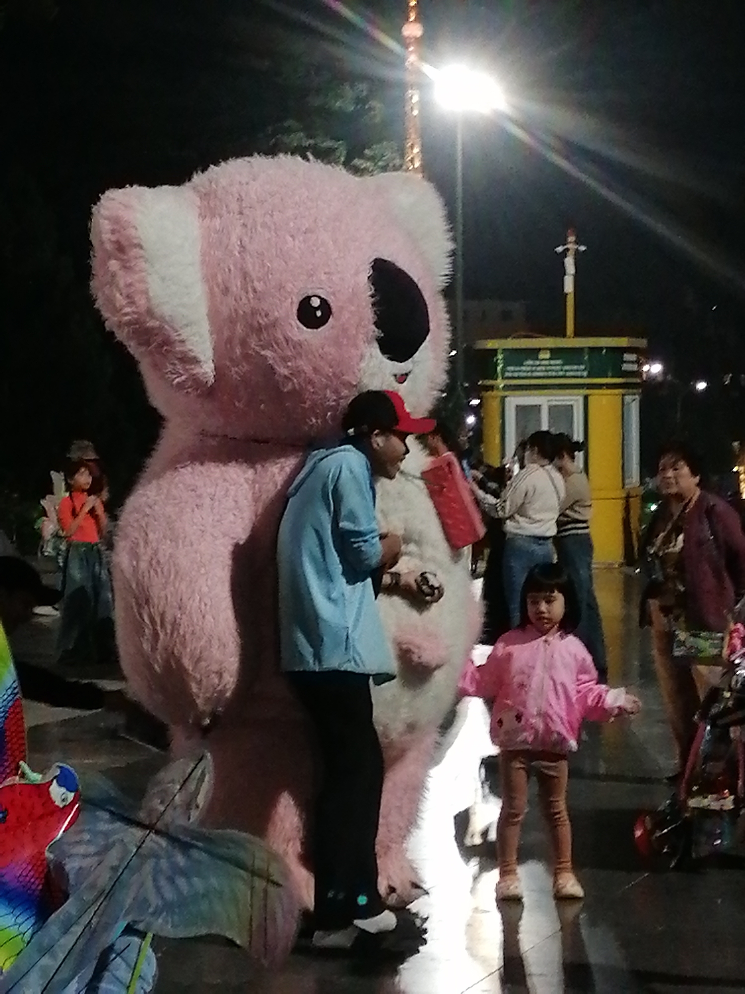 The image depicts an outdoor scene at night, possibly in Da Lat, Vietnam, as suggested by the overall atmosphere and potential local architecture. A very large, pink, plush koala mascot is the central subject. To its right, a person in a blue jacket and black pants stands next to the mascot, appearing to be interacting with it or posing. In front of the mascot and to its left, a young child in a pink jacket and brown leggings stands facing forward. Behind the koala, several other individuals are visible, including a woman in a dark jacket and a woman in a light-colored top. A yellow building with green trim is in the background, and a bright street lamp illuminates the scene, casting reflections on the wet ground. There are also elements of colorful decorations or props in the foreground, including what appears to be part of a kite. The lighting conditions suggest it is evening or night, with the street lamp being a prominent light source.