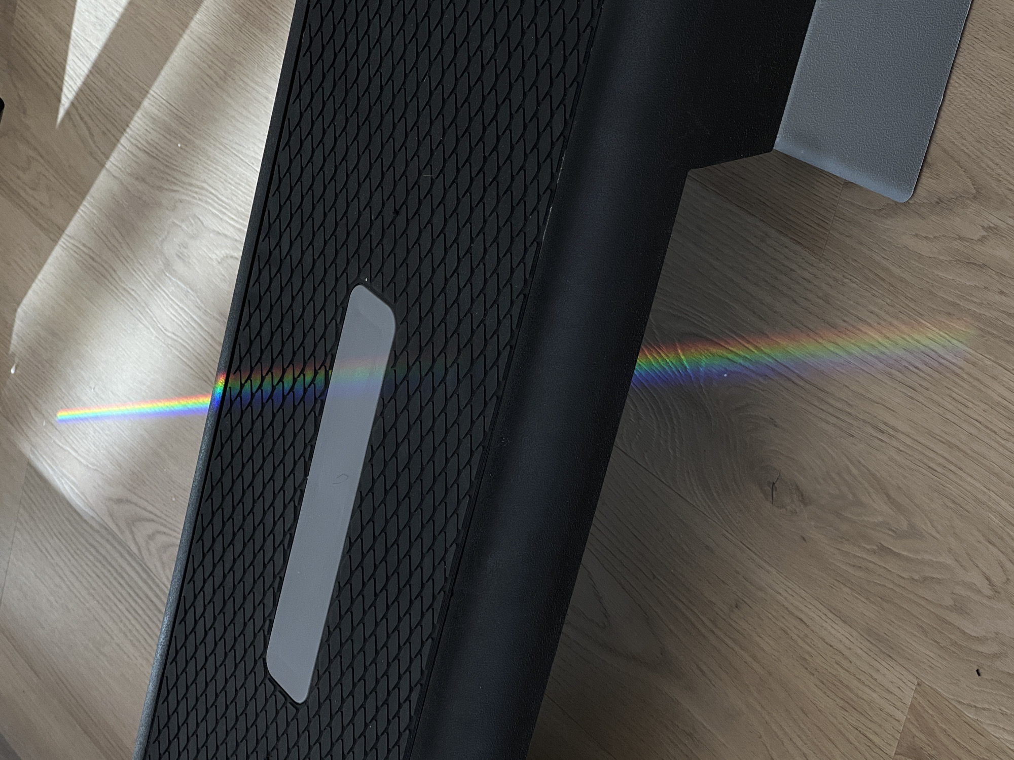 A close-up, high-angle shot displays part of a black, textured surface with a gray stripe on the left side, likely a step or platform. A spectrum of rainbow light spans across the step and continues onto the wooden-textured flooring below and to the right of it. The location appears to be indoors, and there is no indication of the image being taken in Budva, Montenegro. No people or actions are visible.