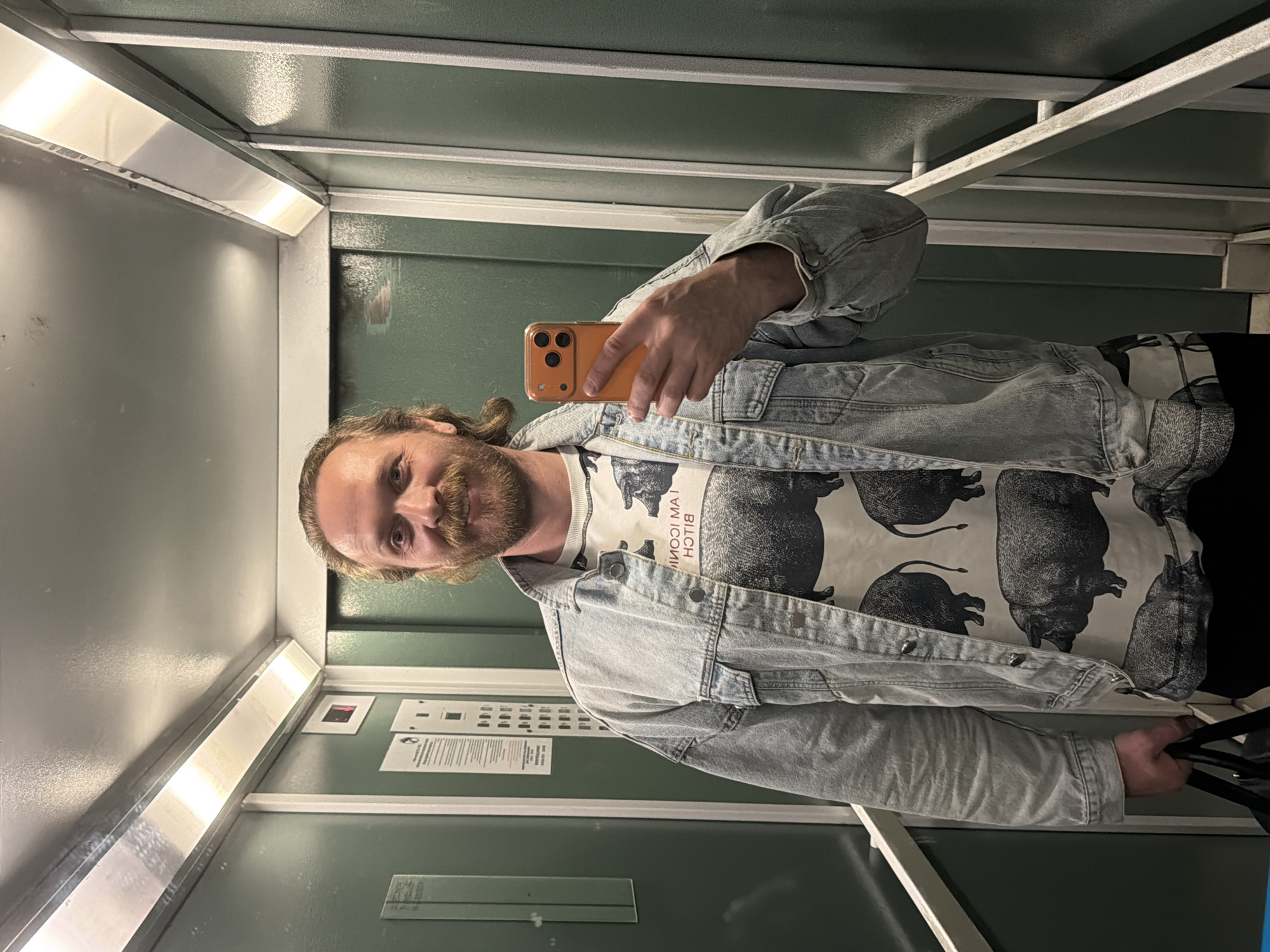 A man is taking a selfie in an elevator. He is holding an orange smartphone in his right hand. He has long blond hair, a beard, and is wearing a light blue denim jacket over a white t-shirt with a black graphic of a pig. The elevator has green paneled walls and a mirrored side with fluorescent lighting. There are informational plaques and buttons on the wall to the right of the man. The location is likely in Saint Petersburg, Russia, based on typical elevator designs in that region.