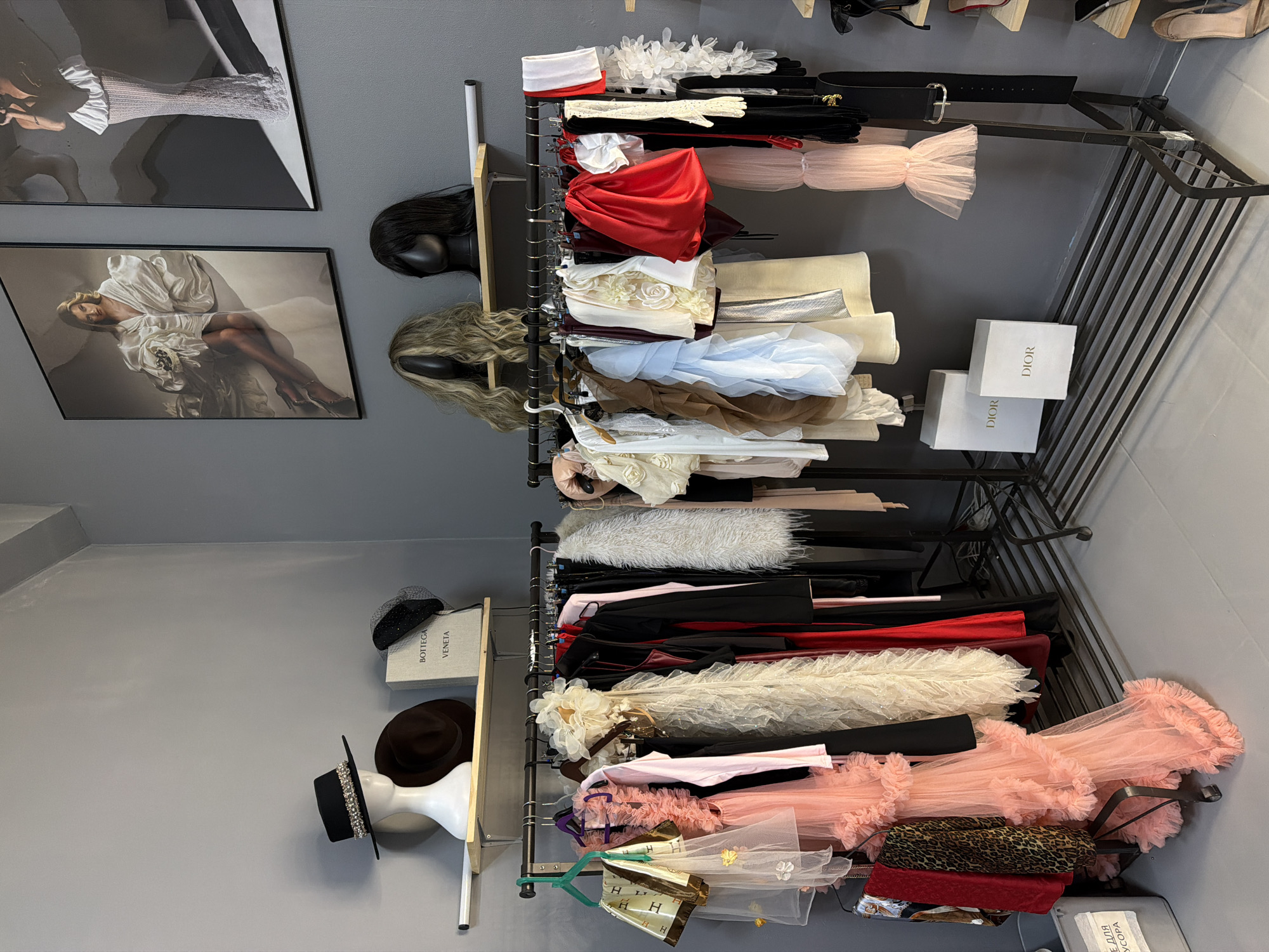 The image shows a room in Kazan, Russia, with a rack of clothing and accessories. The walls are gray. Two framed fashion photographs are hanging on the left wall above two wig stands. To the right of the wig stands is a clothing rack holding various garments, gloves, and a black belt. Two Dior boxes are on the rack. On the left wall is a shelf holding a black hat and books. The floor is not visible. The room appears to be a closet, shop, or storage area for clothing and fashion items.