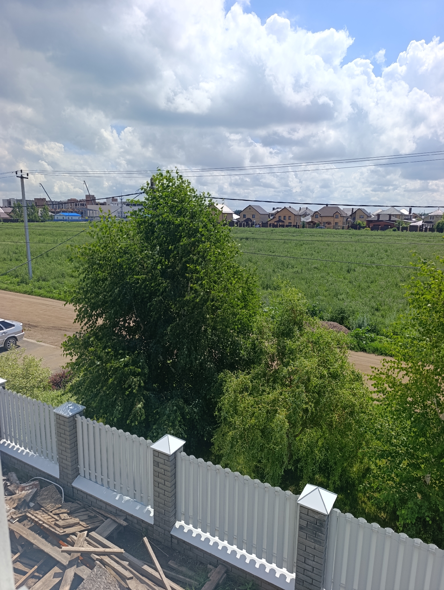 The image shows a view from an elevated position, looking out over a fence and into a suburban or rural landscape. In the foreground, there is a white picket fence with brick pillars. Below the fence, there is a pile of construction debris, consisting of wooden planks and other building materials.
Beyond the fence, a dirt road is visible, and a silver car is parked on its edge. Lush green trees are prominent in the middle ground, with one large, leafy tree dominating the left side of the image. Further back, a vast green field stretches out, dotted with scattered wildflowers. In the distance, a row of houses with dark roofs forms a suburban skyline, and behind them, a few multi-story buildings under construction with cranes are visible. The sky is a mix of blue sky and fluffy white clouds. The scene suggests a quiet day in a developing residential area, possibly on the outskirts of a town.
The metadata indicates the image was taken in Ivanovka, Russia.