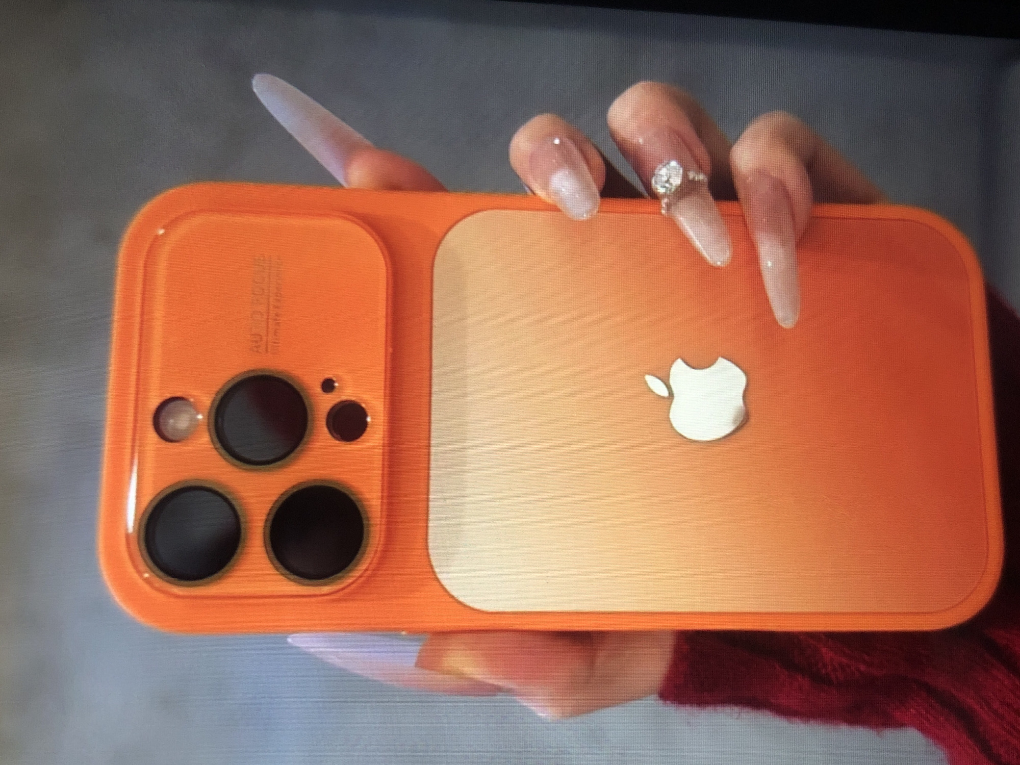 An orange cell phone with an Apple logo is being held by a person. The person has long, light-colored fingernails, one of which has a ring on it. The phone case has three camera lenses on the left side. The background is a blurred gray. The location is not discernible from the image.
