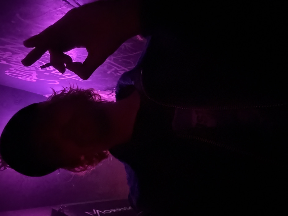 A person is lit with a purple light, possibly from below. Their hand holds a cigarette, fingers extended. They are wearing a dark jacket with a zipper. The back of their head shows curly hair. There is graffiti on the wall behind the person. The word "MORRIS" is visible on what appears to be a keyboard case.