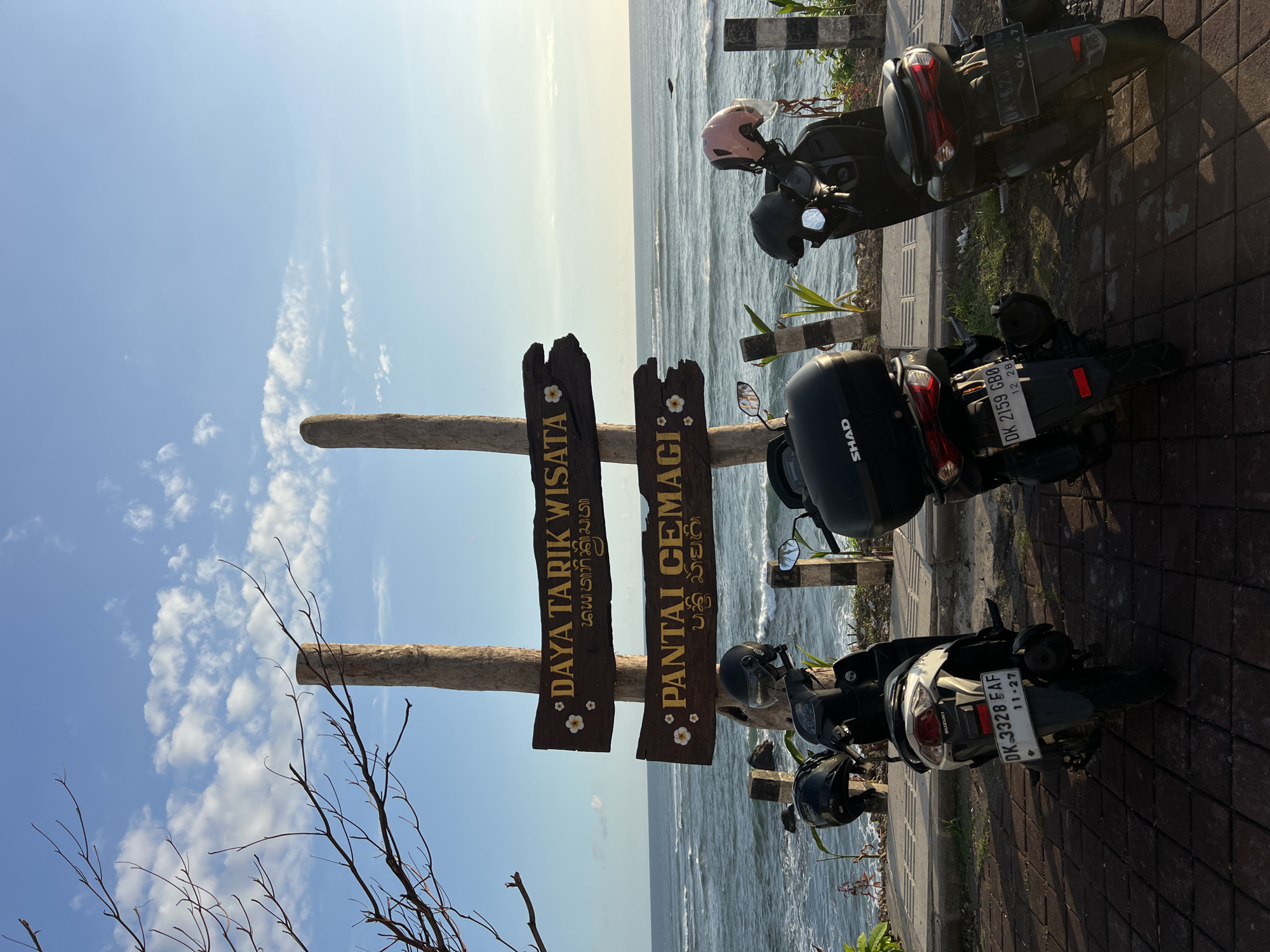 The image shows a coastal scene in Mengwi, Indonesia. Two wooden signs read "Daya Tarik Wisata" and "Pantai Cemagi." Several parked scooters line a paved walkway, facing the ocean. License plates read "DK 3328 EAF," "DK 2159 GBO," and "DK-6273 NG." Helmets rest on the scooters. The ocean is visible in the background under a partly cloudy sky.