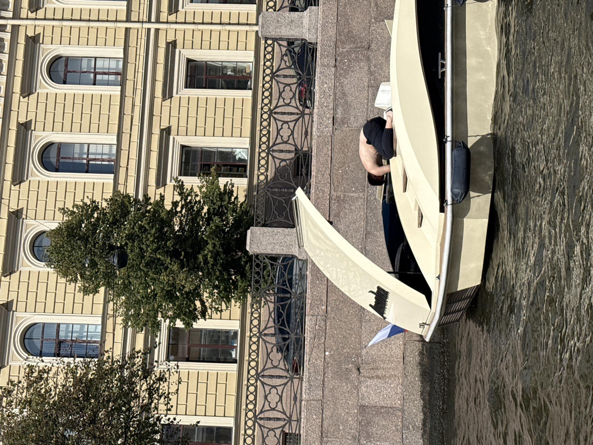 A person is kneeling on the deck of a light-colored boat, with their torso facing downwards and head obscured as they appear to be working on something. The boat is on a body of water, likely a canal or river, with a stone embankment to the right. Behind the embankment, a multi-story building with yellow facade and arched windows is visible, suggesting an urban setting in Saint Petersburg, Russia. A Russian flag is attached to the back of the boat. The weather appears to be sunny.